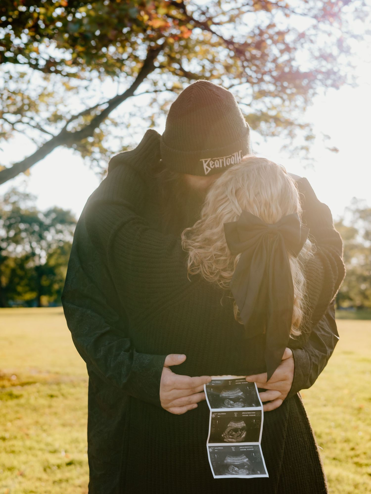 Couple sharing intimate moment while holding sonogram pictures in golden sunlight.