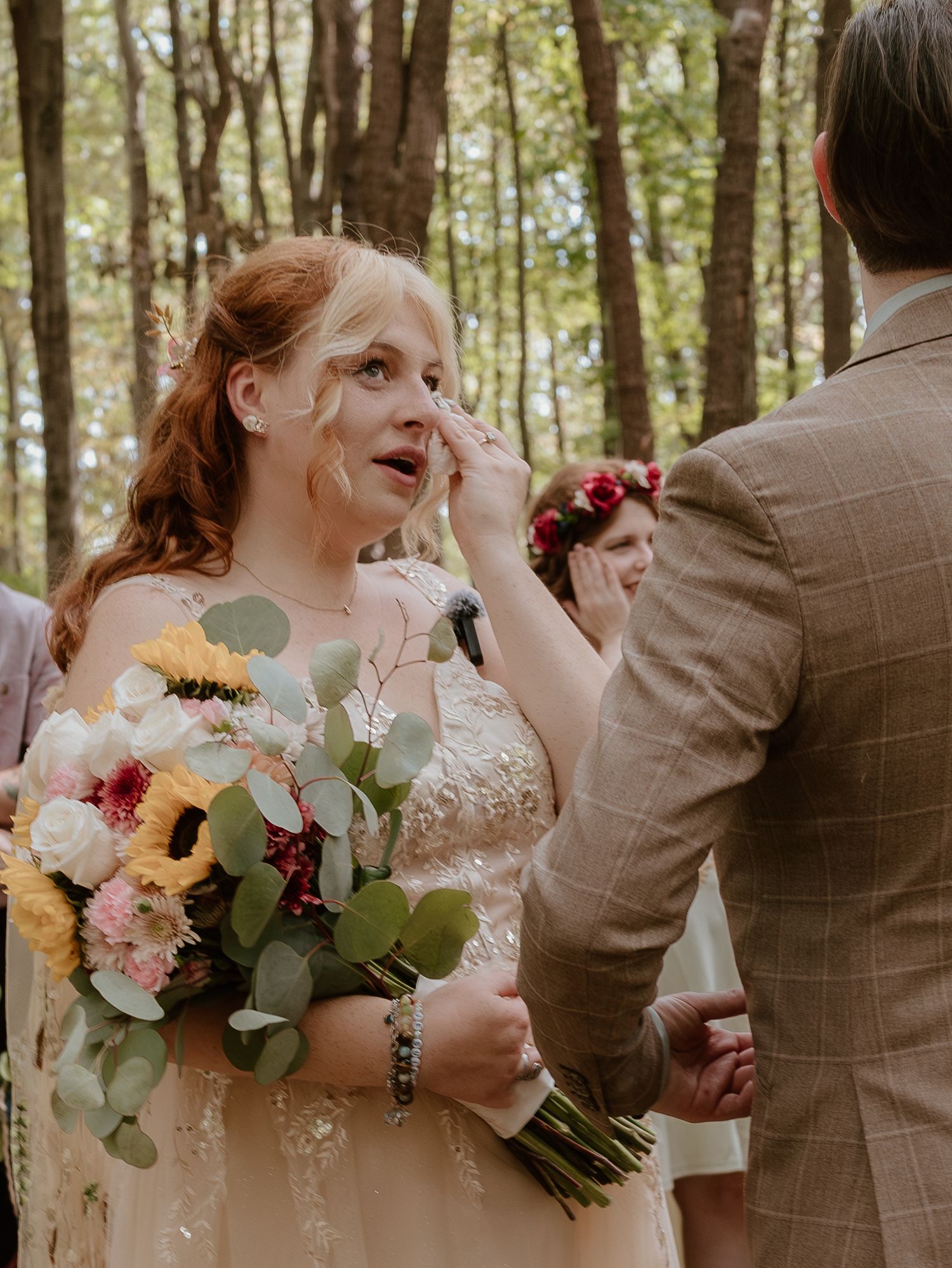 Wedding party members hold colorful bouquets with sunflowers and pink flowers while laughing in a wooded outdoor setting.