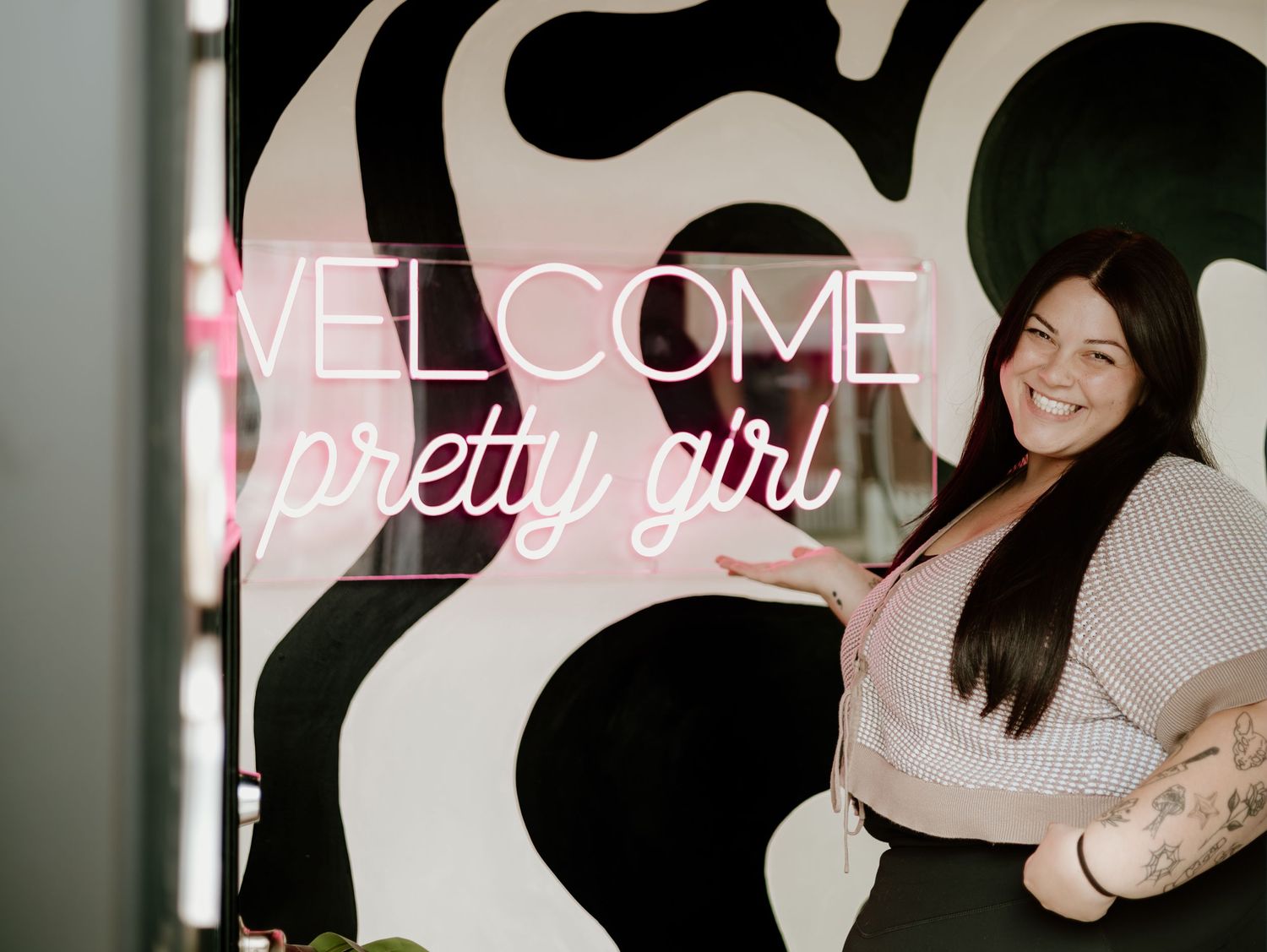 Neon pink 'Welcome pretty girl' sign glows against black and white swirled wall beside person in beige sweater.