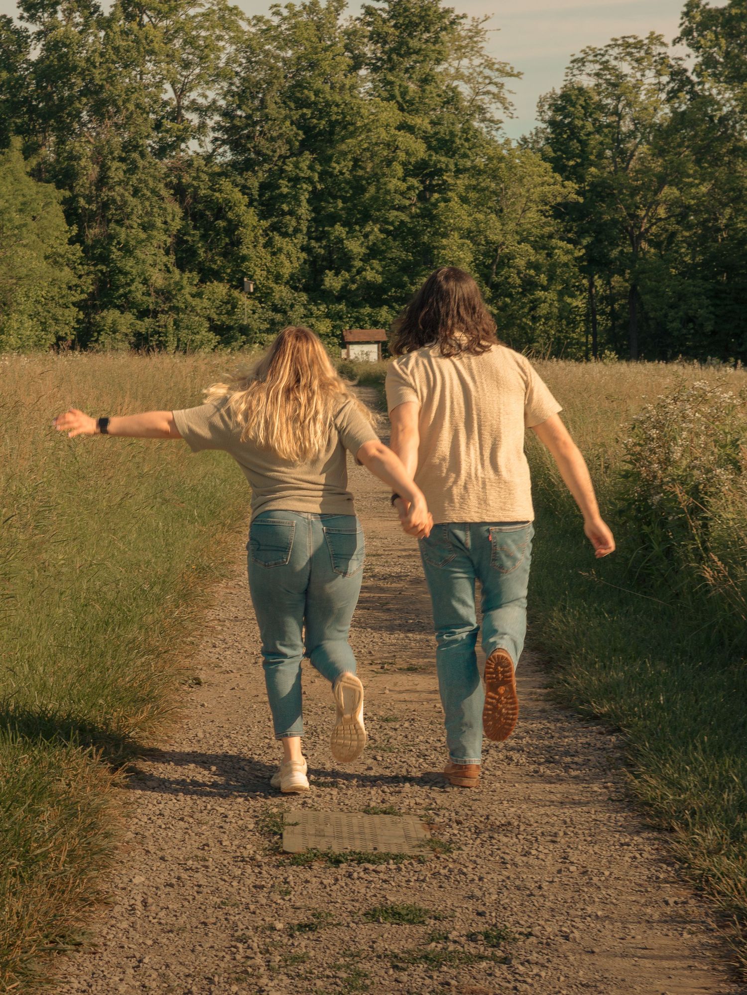 Two people in jeans and light tops walking together on a dirt path through a sunny meadow on a summer day.