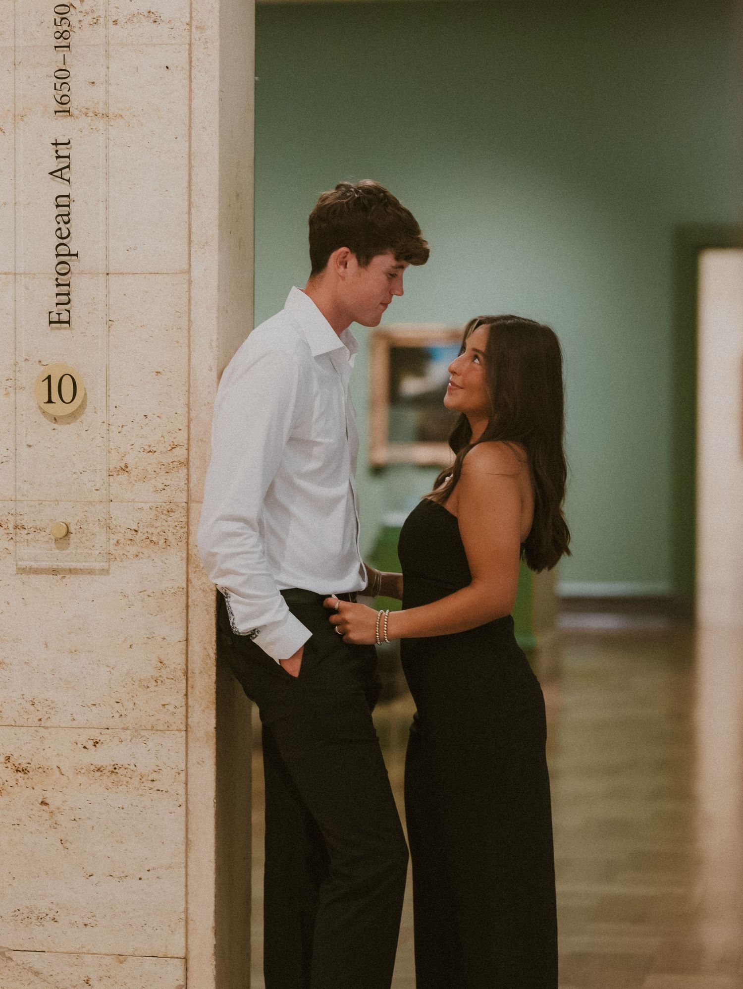A couple in formal attire shares an intimate moment in a hallway with white stone walls and minimalist decor.