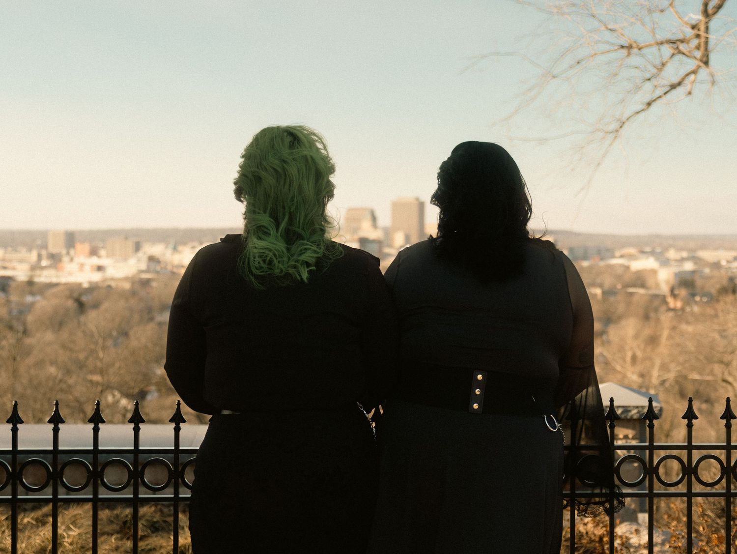Silhouettes of people standing against an iron fence at sunset in a cemetery with golden light filtering through.