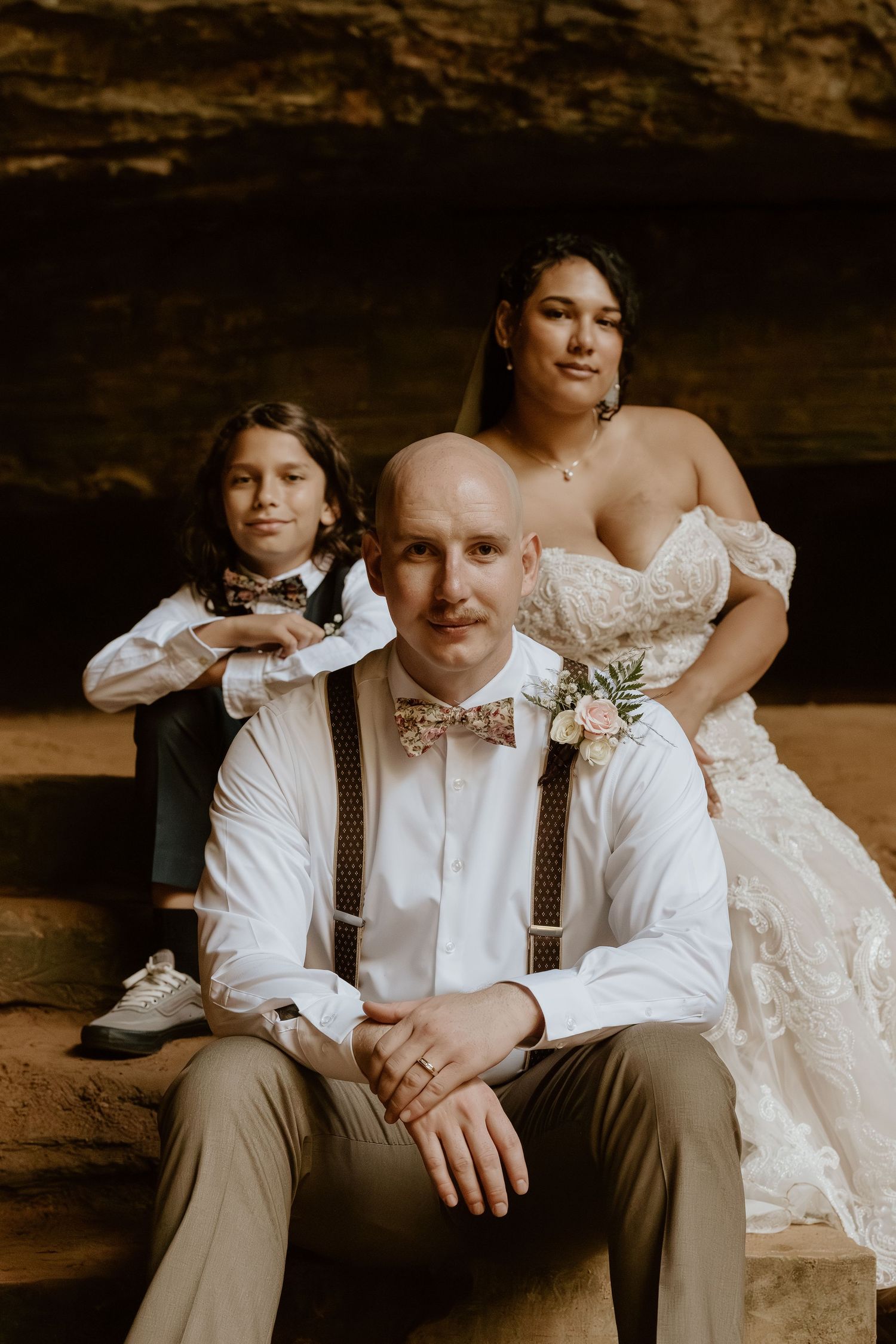 Wedding couple in rustic setting with groom in suspenders and bride in lace dress sitting close together.