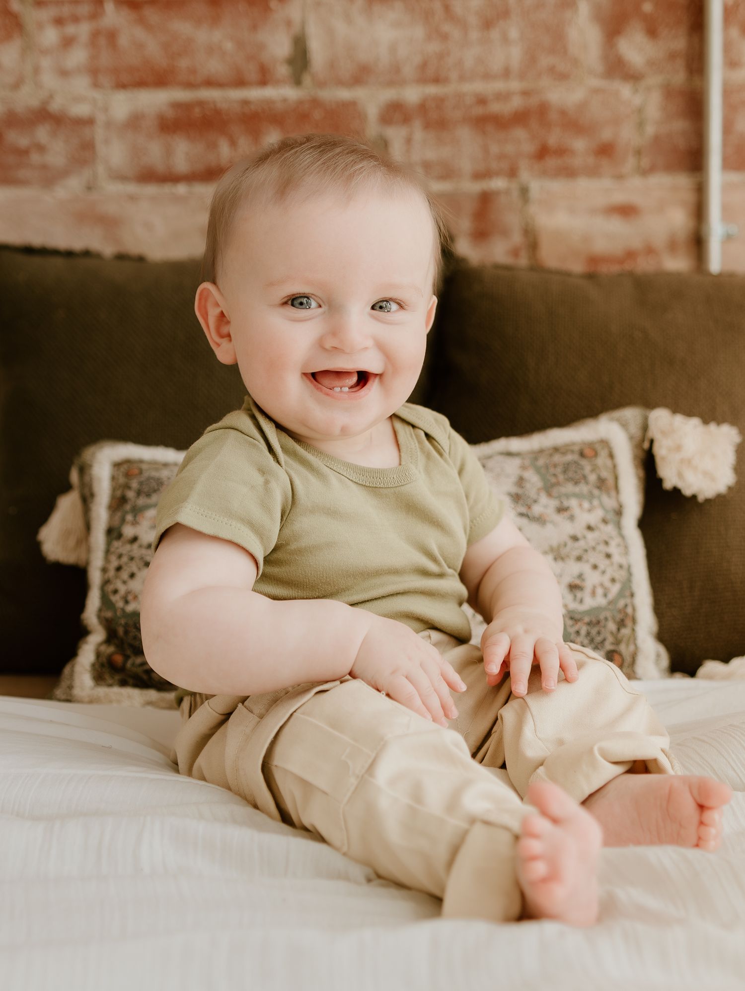 A cheerful baby wearing a beige outfit sits on a brown couch against an exposed brick wall.