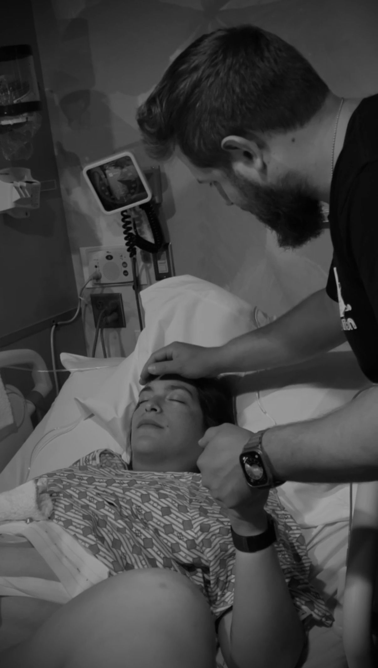 A medical professional examines a patient lying in a hospital bed, with monitoring equipment visible in the background.