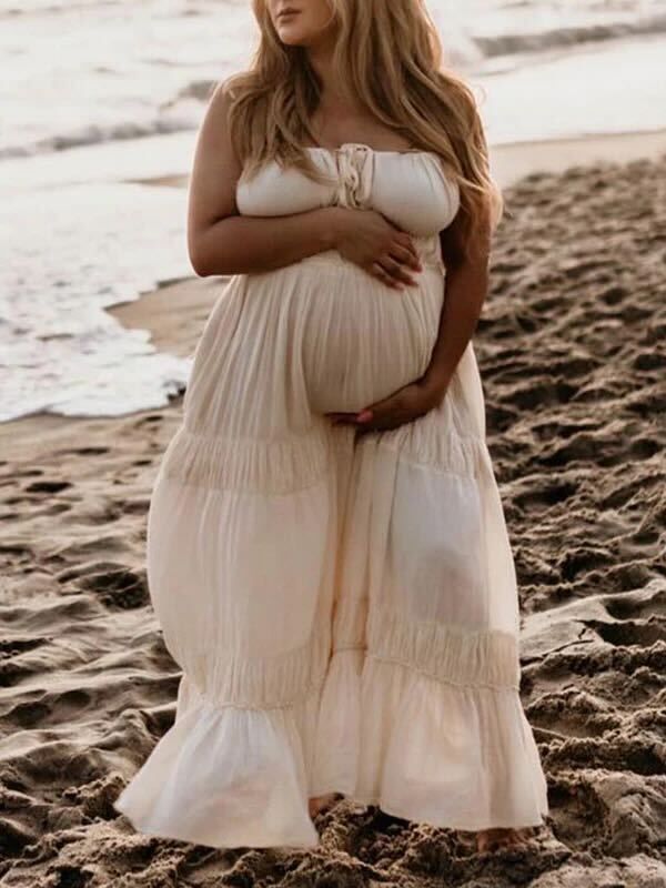 Expectant woman in white flowing tiered maxi dress poses gracefully on beach at sunset.