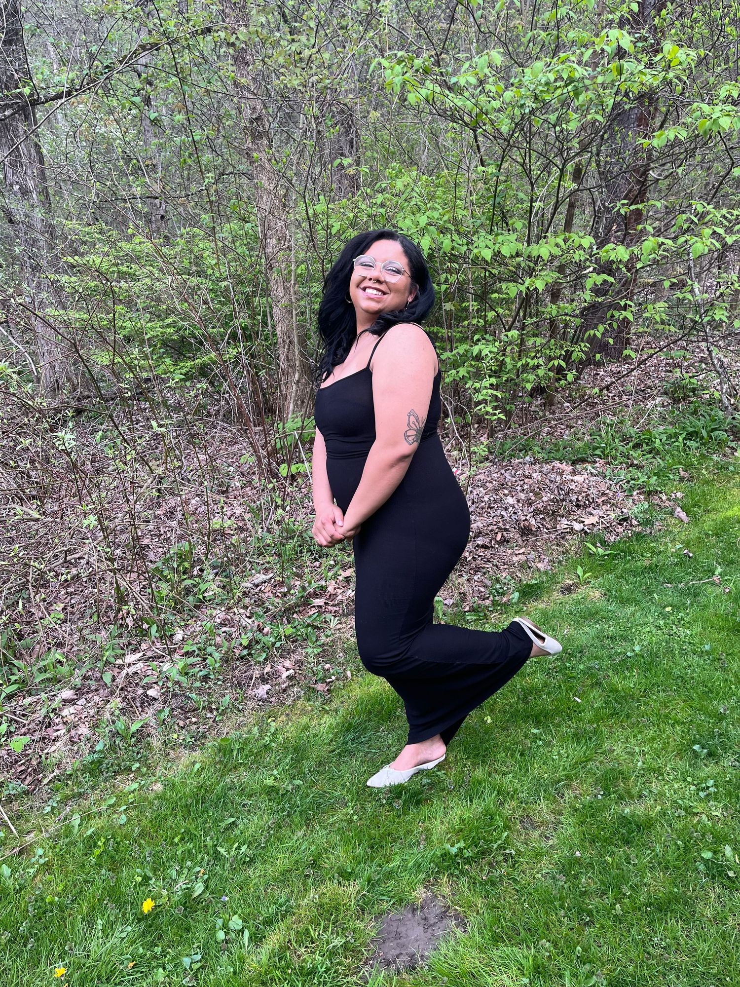 A person in a black outfit posing playfully on grass with trees in the background.