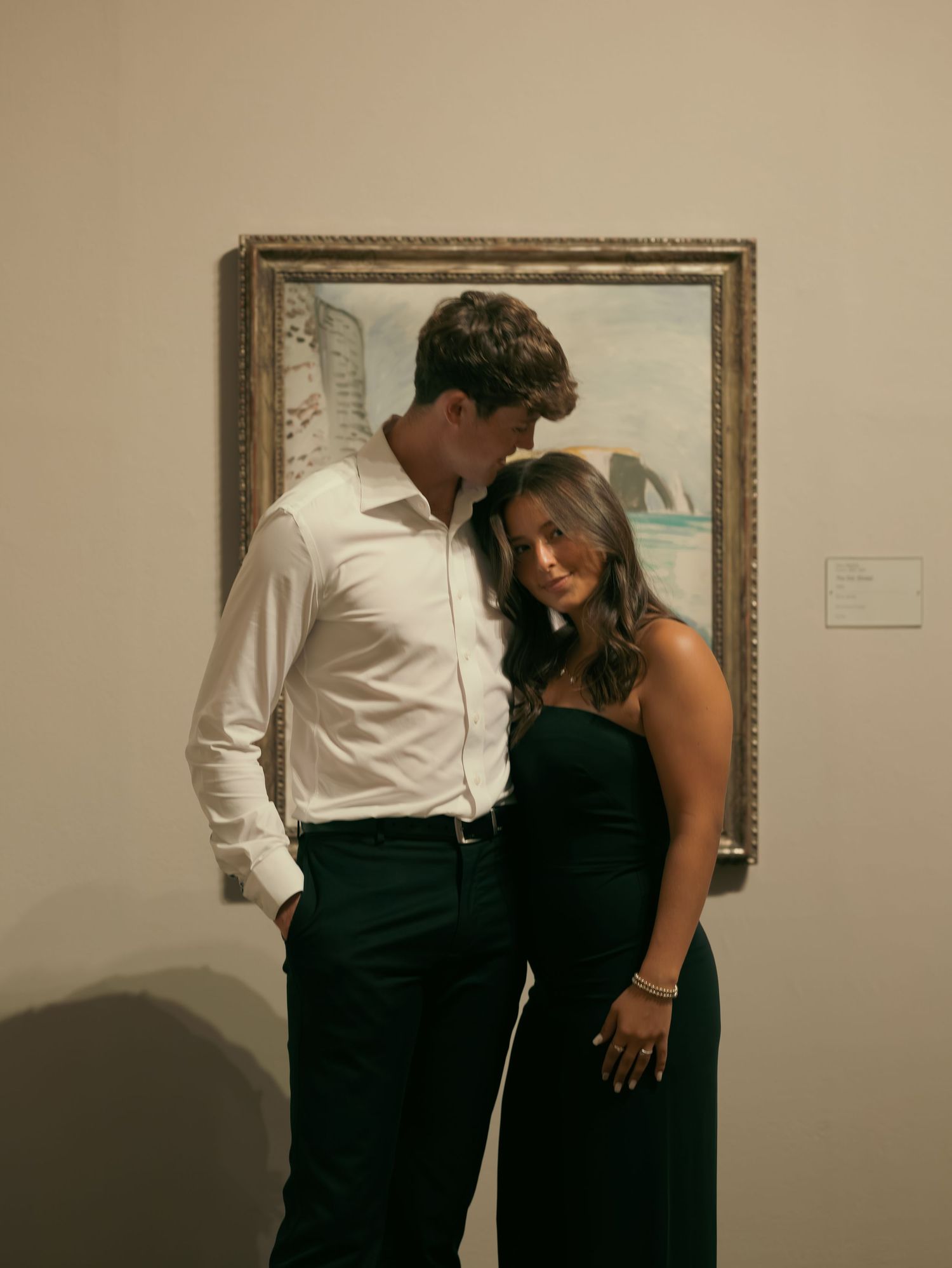 A couple in formal attire share an intimate moment in what appears to be an art gallery or museum space.