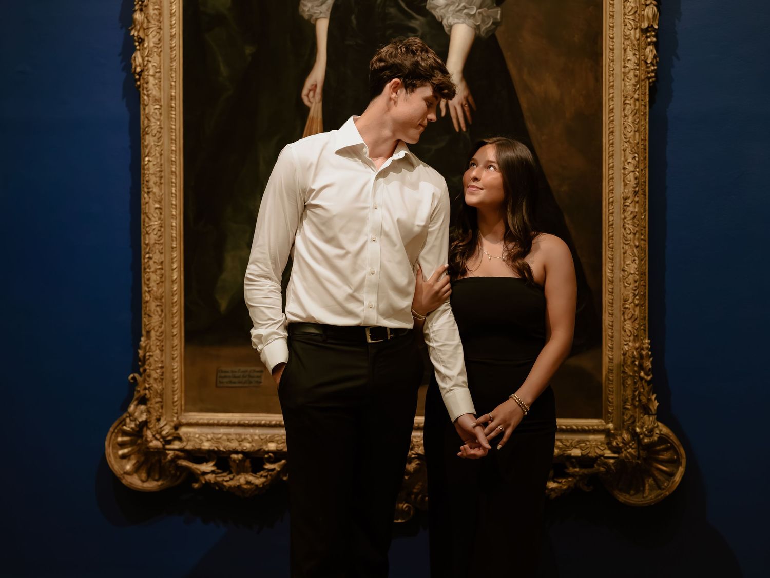 A couple in formal attire shares an intimate moment against a dark blue wall with an ornate gold frame.