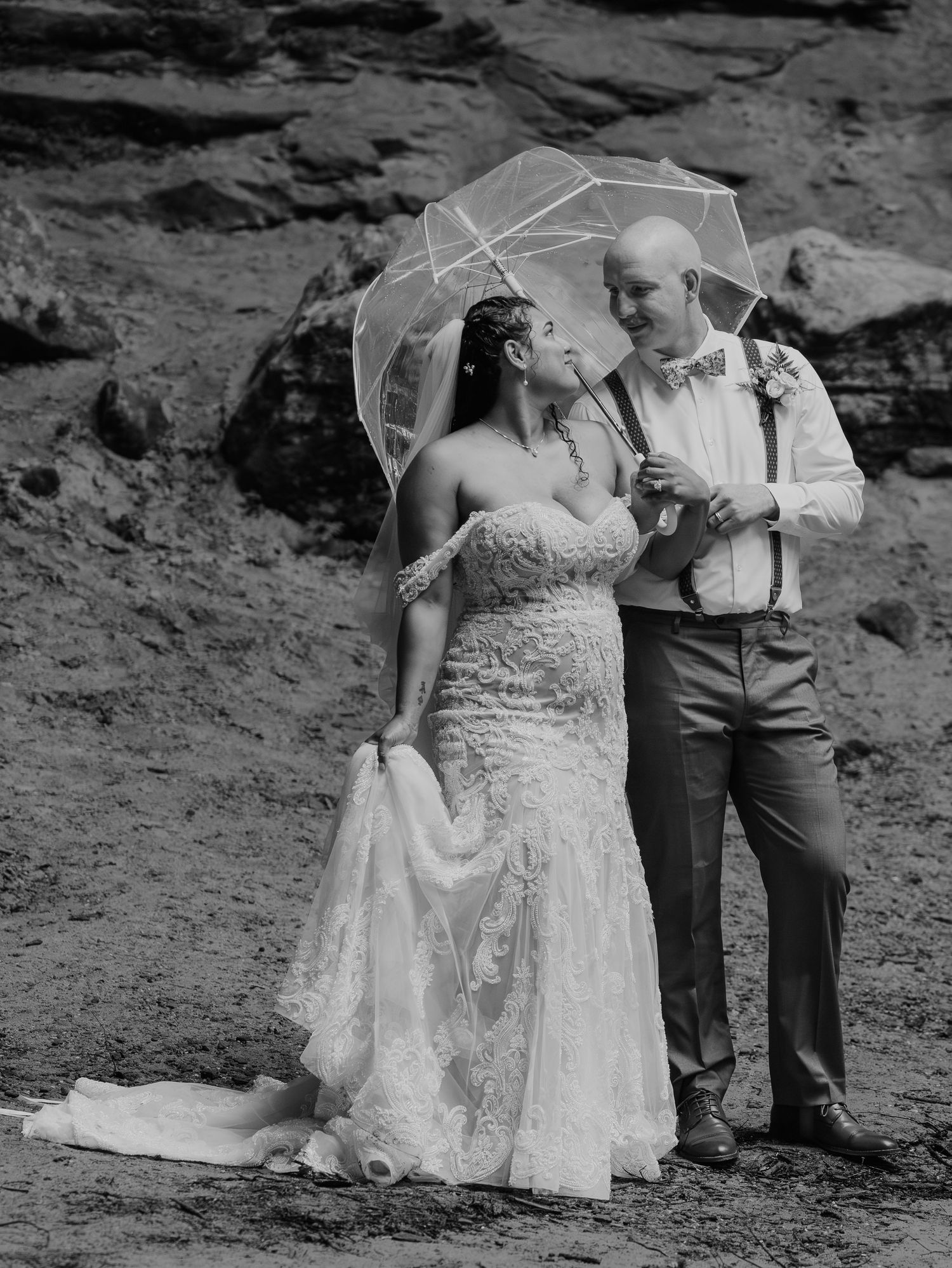 Black and white wedding photo of a couple on rocky terrain, bride in lace dress and groom in suspenders.