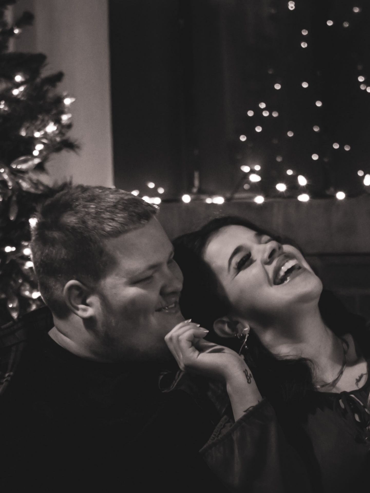 Couple shares a joyful moment by Christmas tree with twinkling lights in black and white photo.
