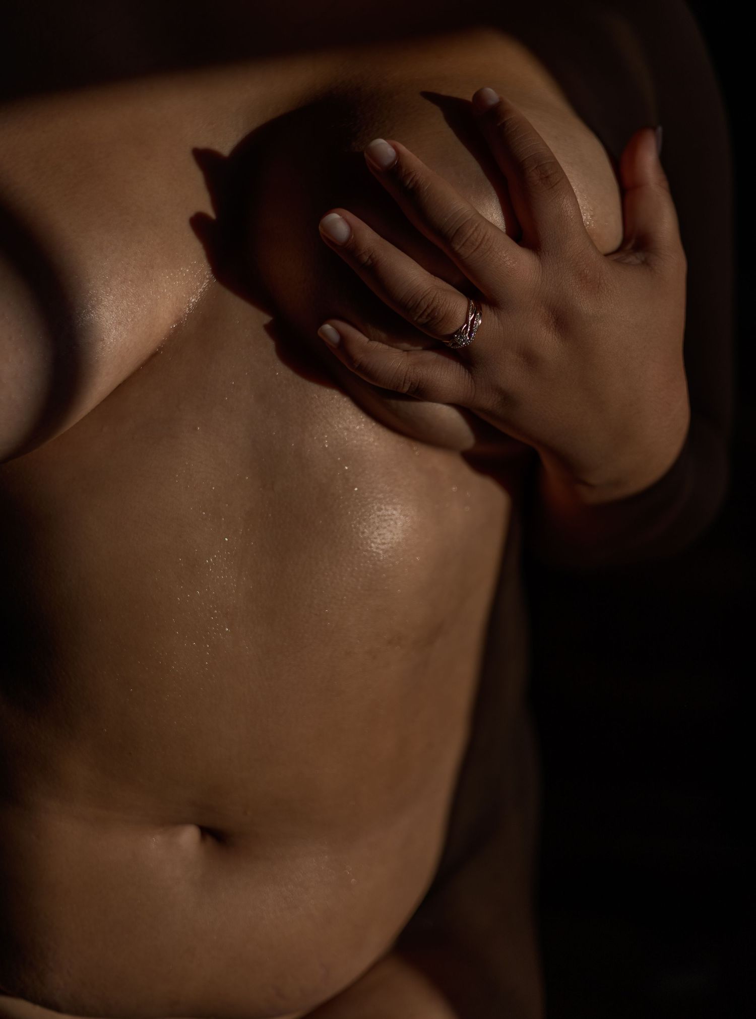 Artistic boudoir photo showing close-up torso and hand with ring against dark background in warm moody lighting.