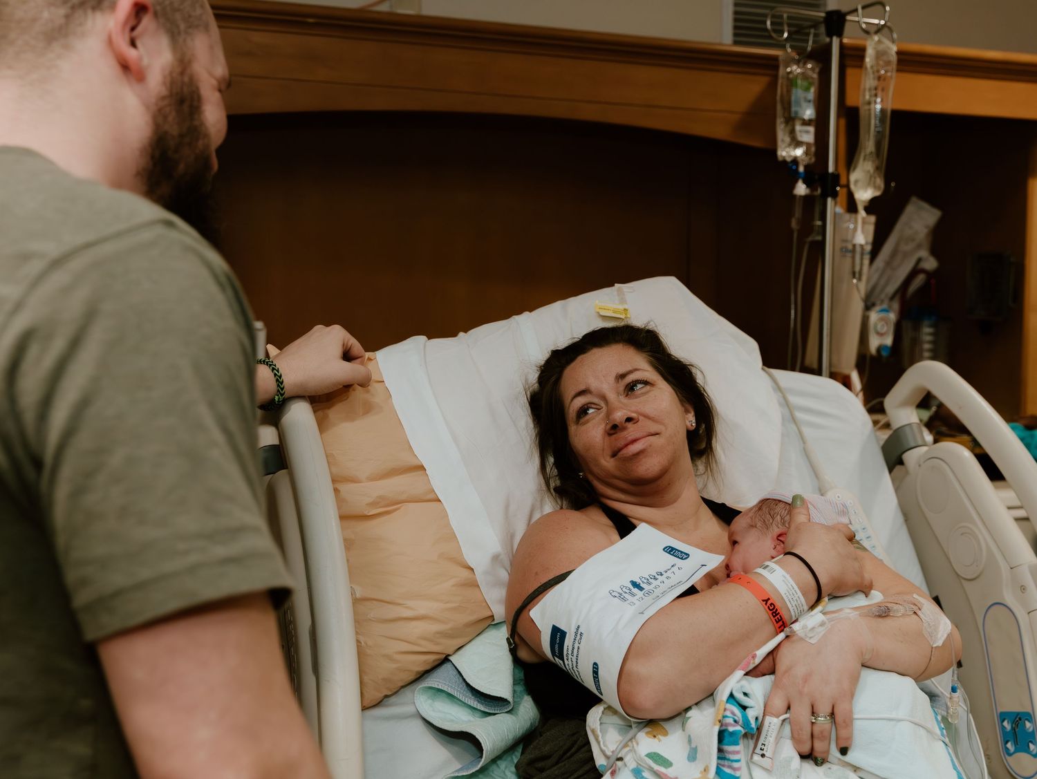 A woman rests in a hospital bed while a man stands beside her in a medical room.