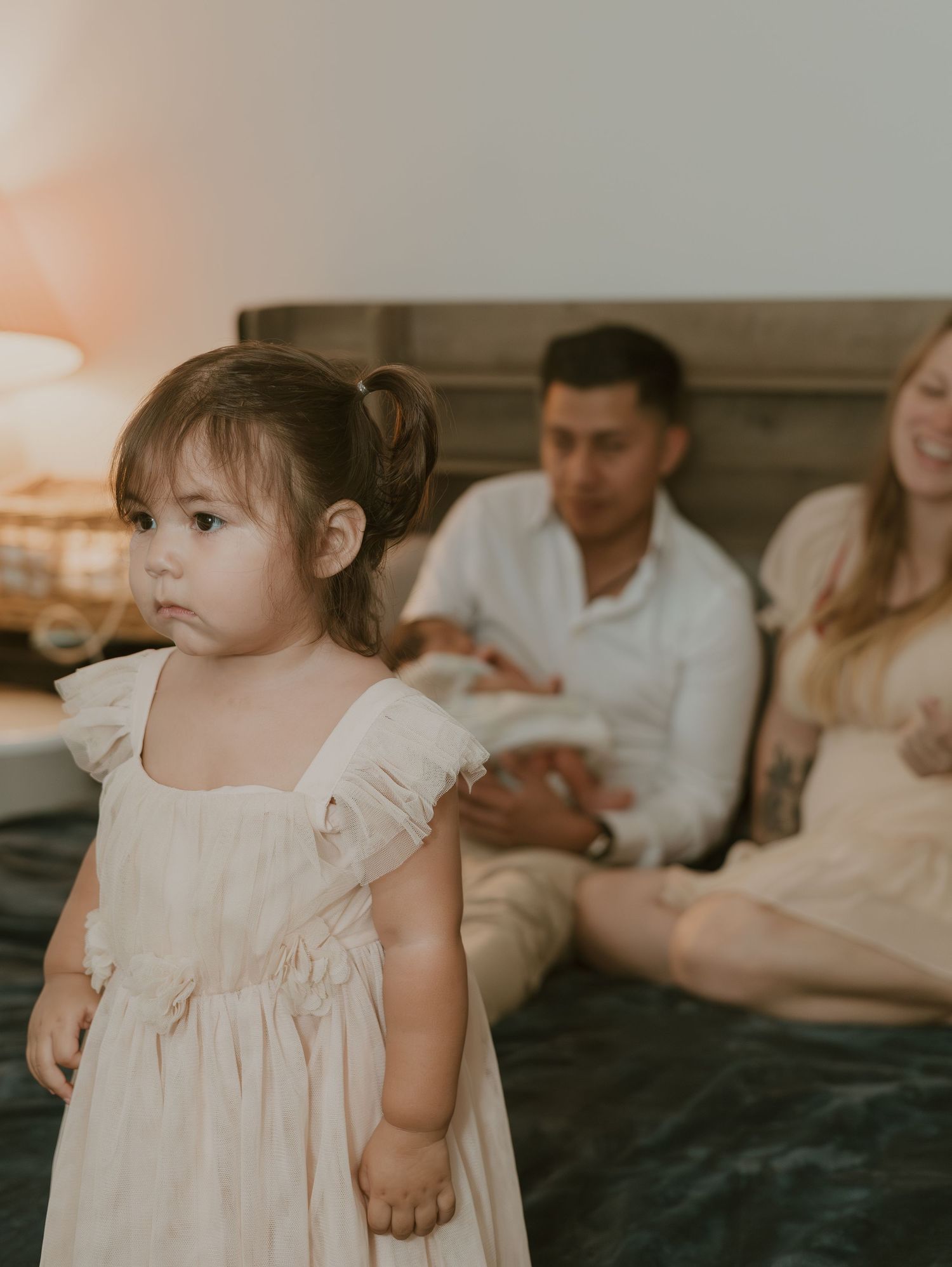 A young child wearing a white ruffled dress stands in the foreground while adults sit on a couch in the background.