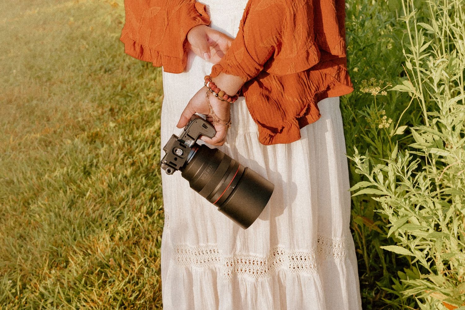 Person in orange jacket and white dress carries leather bag while standing in sunlit grassy outdoor setting.