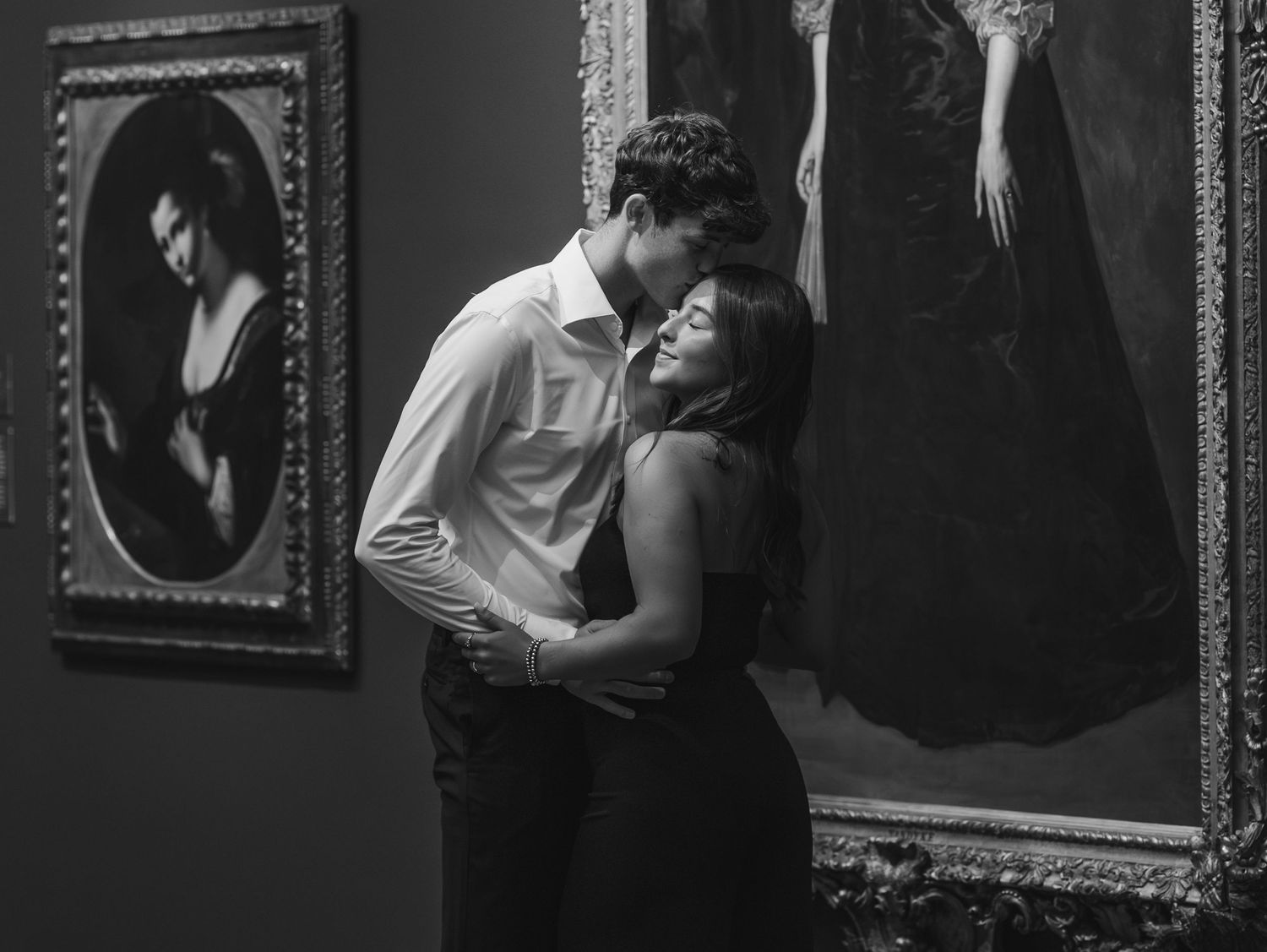 A black and white photograph of people viewing artwork in a gallery with ornate frames on dark walls.