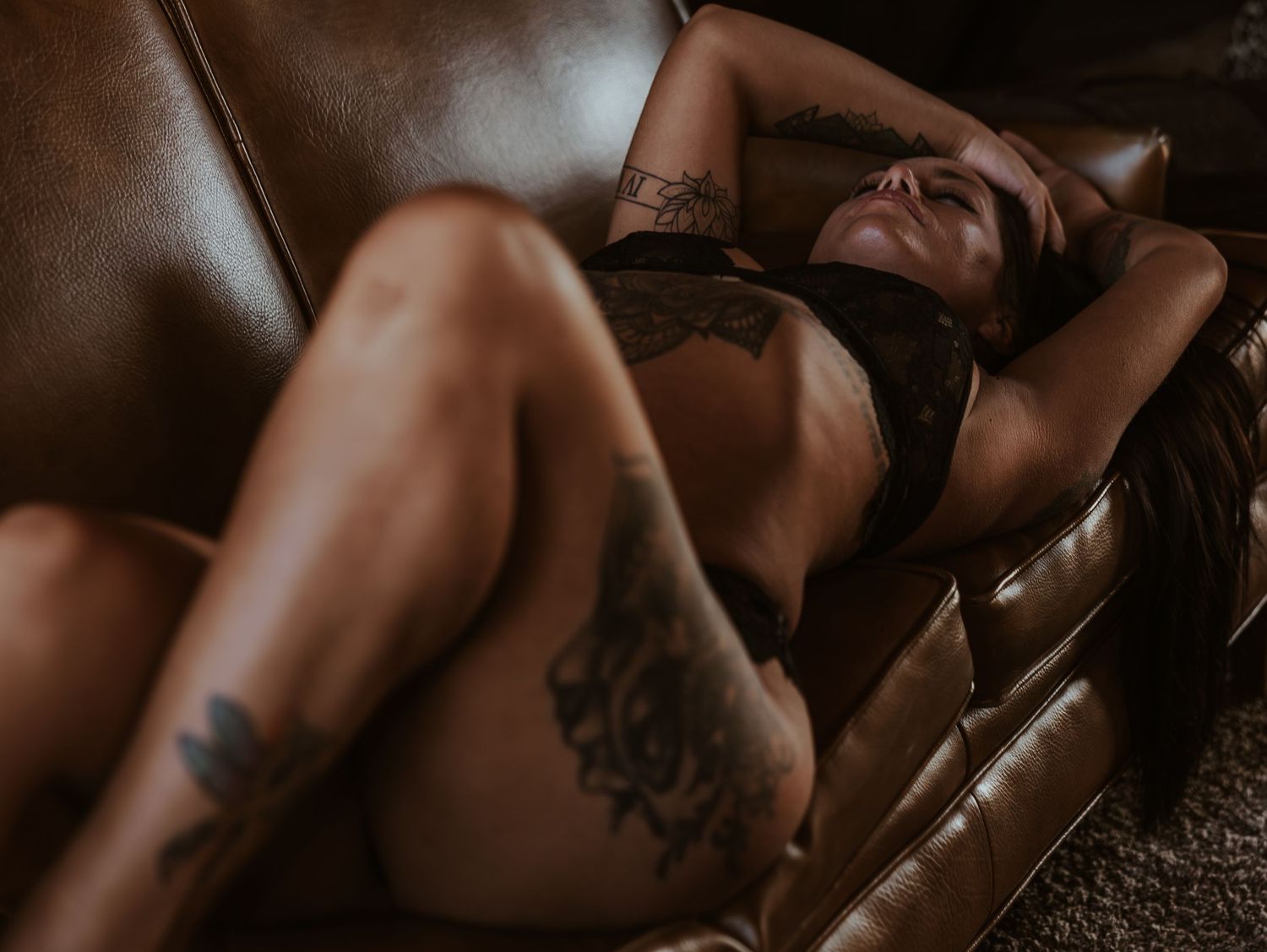 Artistic boudoir photo of person reclining on brown leather couch in intimate lingerie with visible tattoos.