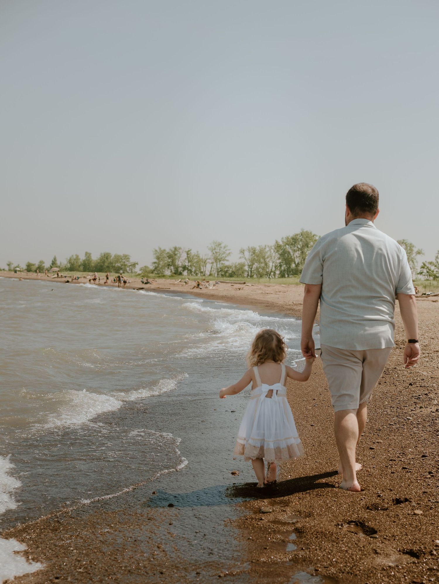 Two people stand at the edge of a rocky beach looking out at the waves and palm trees along the shoreline.