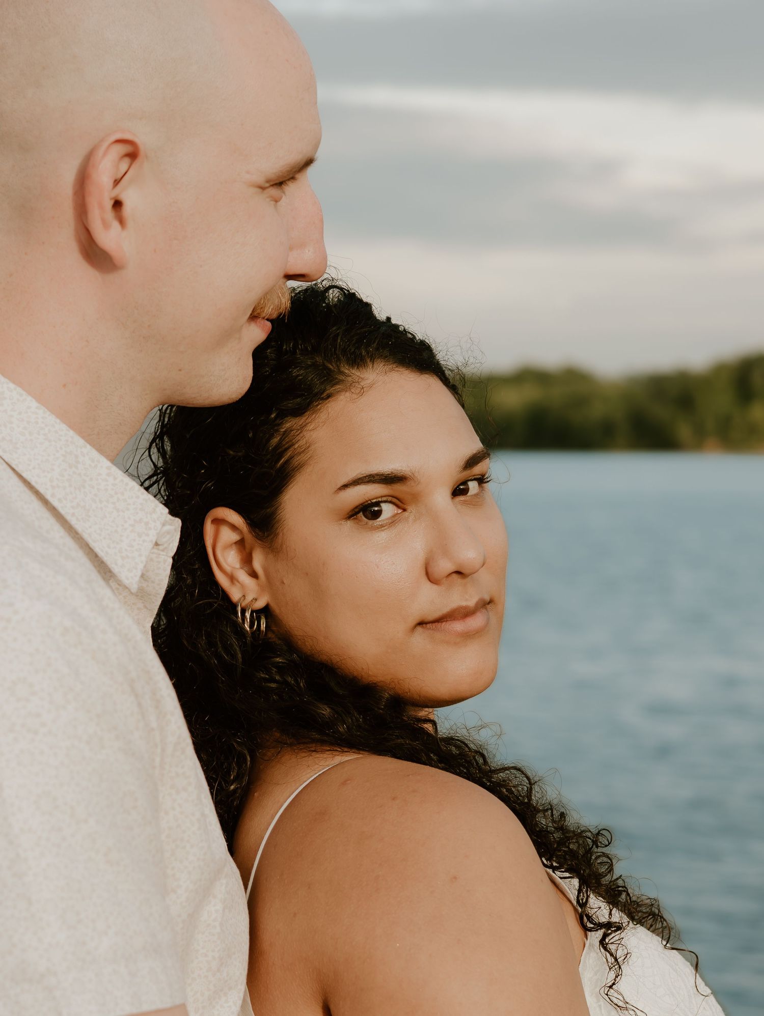 A couple shares an intimate moment by a serene lakeside setting during a wedding photoshoot at sunset.