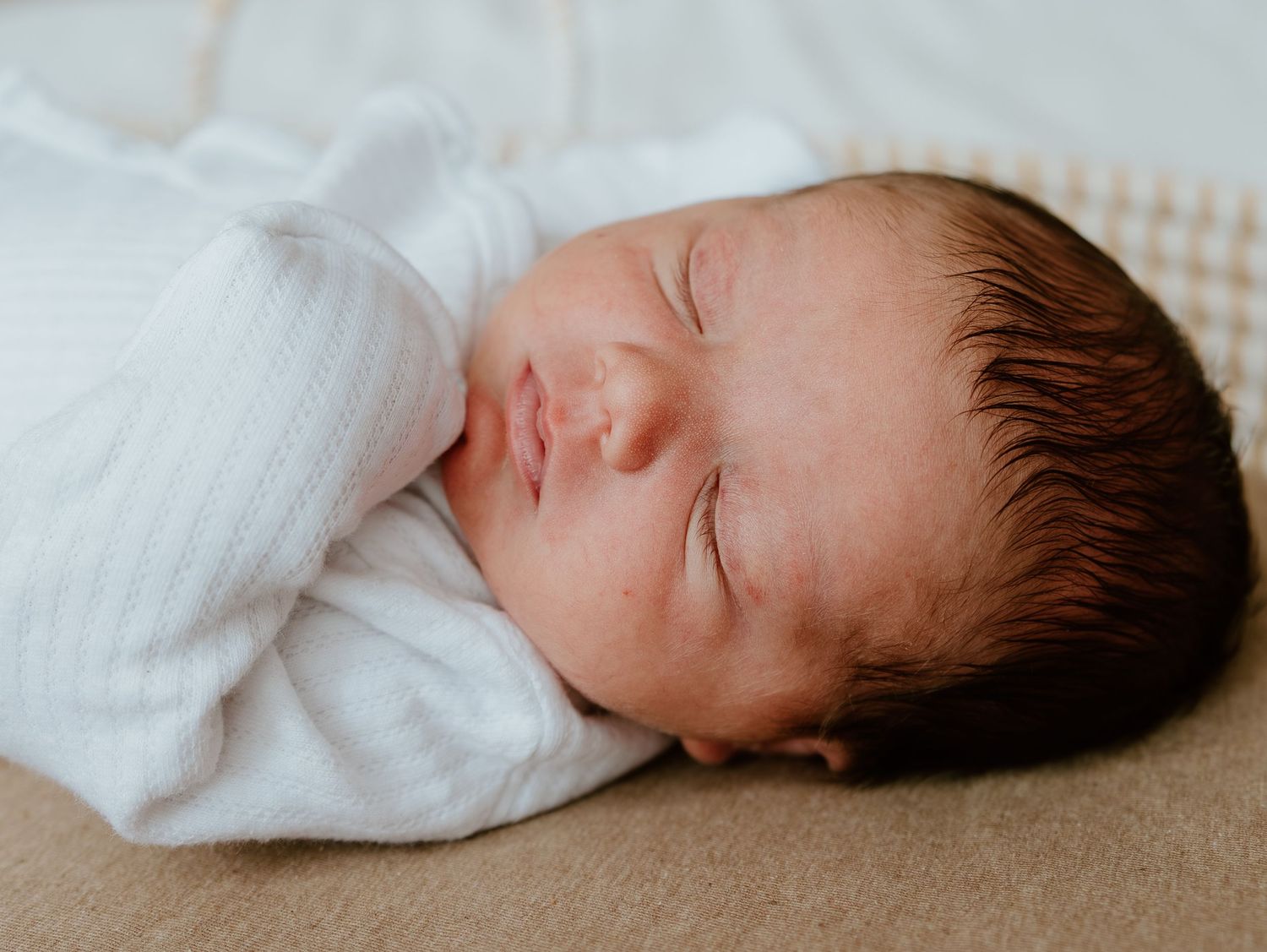 A peaceful sleeping newborn baby wearing a white onesie rests on a beige surface.
