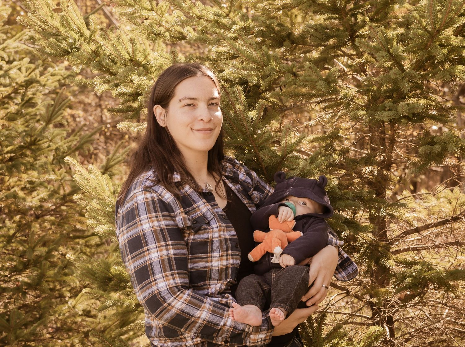 A person in a plaid shirt holds a baby wearing dark clothing with a small orange toy in a wooded outdoor setting.