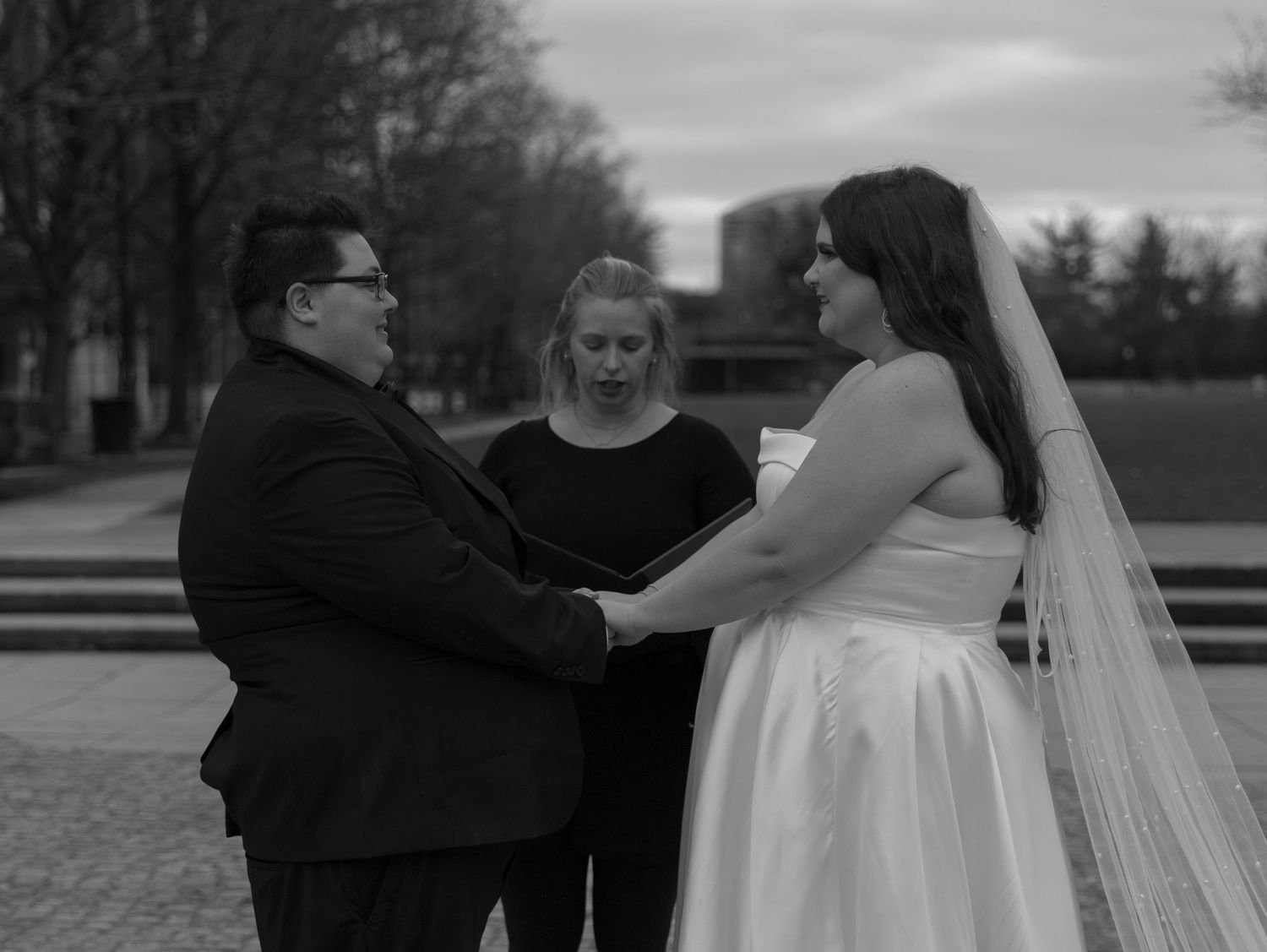 A couple joins hands at their outdoor wedding ceremony as an officiant leads the service in black and white.