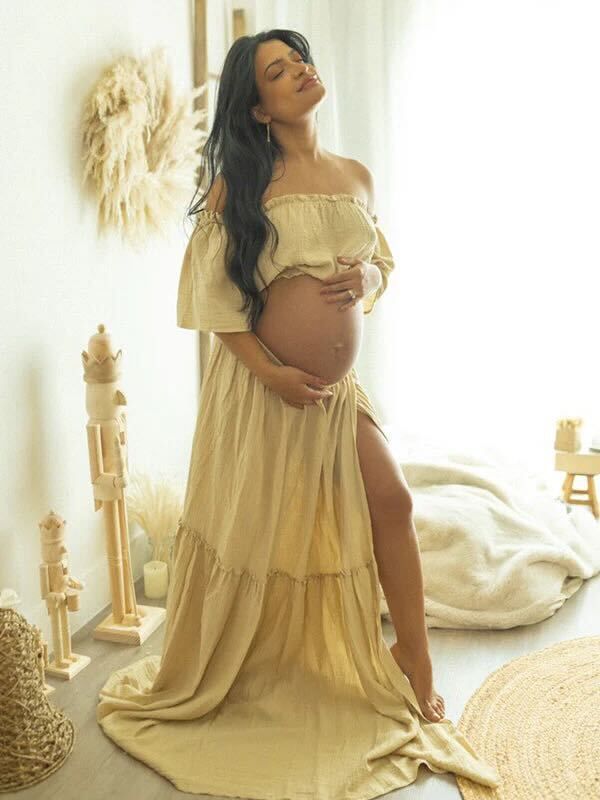 An expectant mother in a flowing off-shoulder cream dress poses by a window with boho decor and natural light.