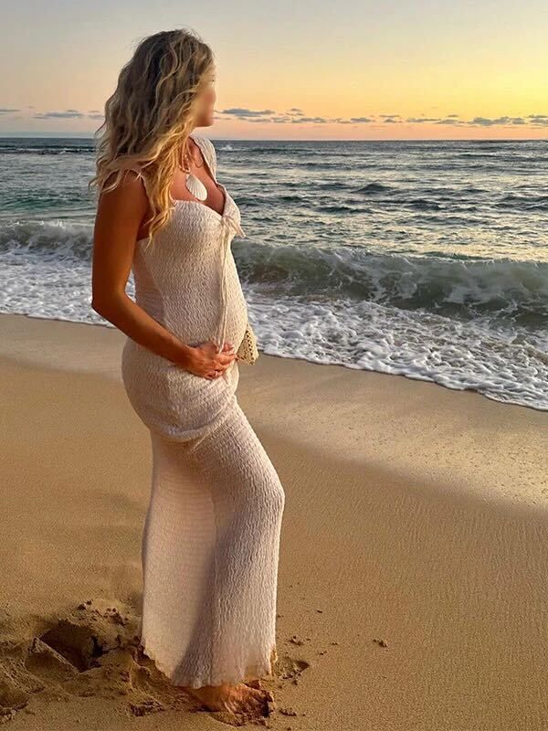 A pregnant woman in a white knit dress stands on a beach at sunset as waves crash on the shore.