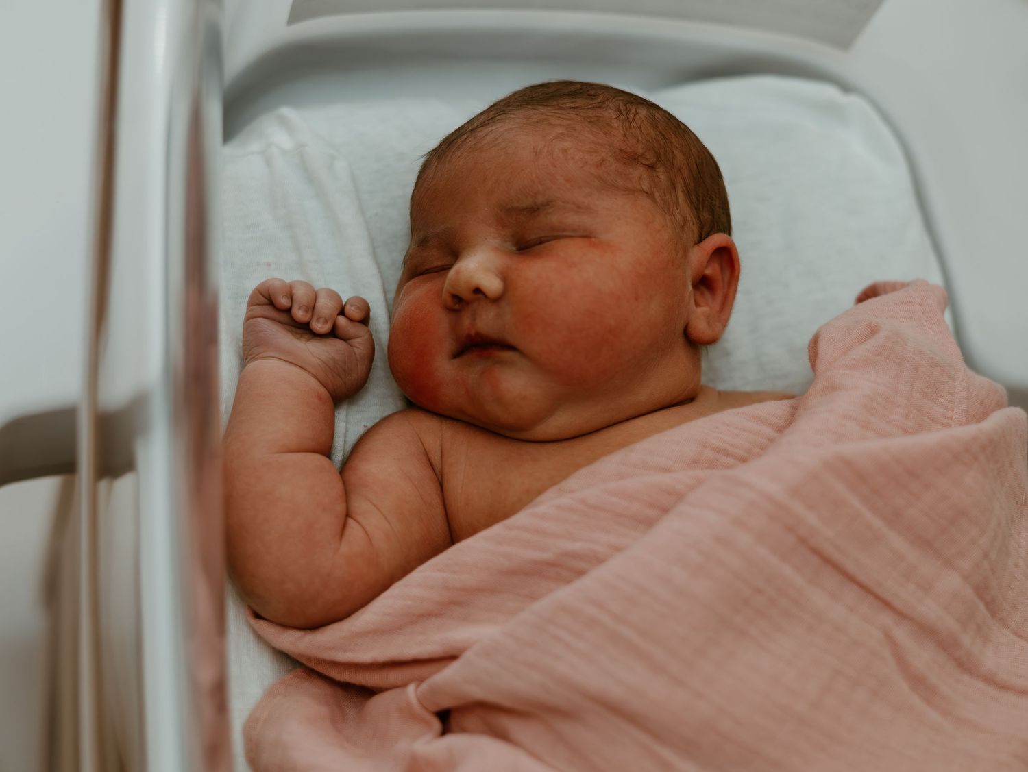 A newborn baby sleeps peacefully in a hospital bassinet wrapped in a soft pink blanket.