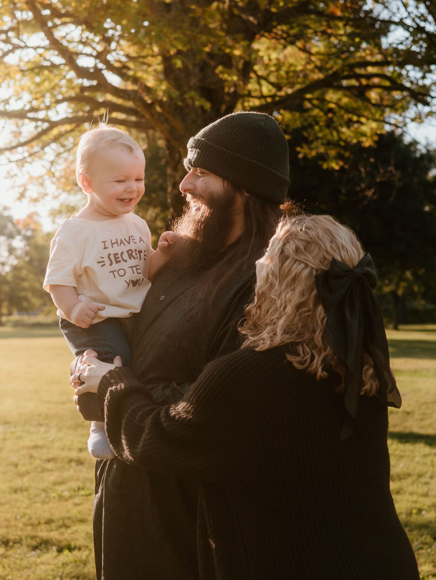 A bearded person holds a small child in golden sunlight during an outdoor autumn photoshoot.