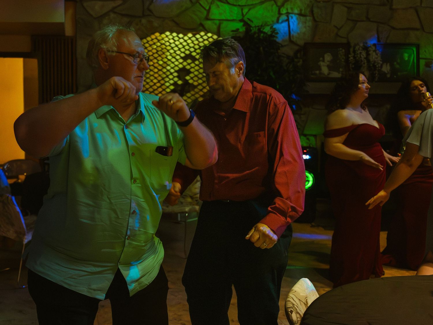 Two men dancing energetically at a party, with guests and decorative lighting visible in the background.