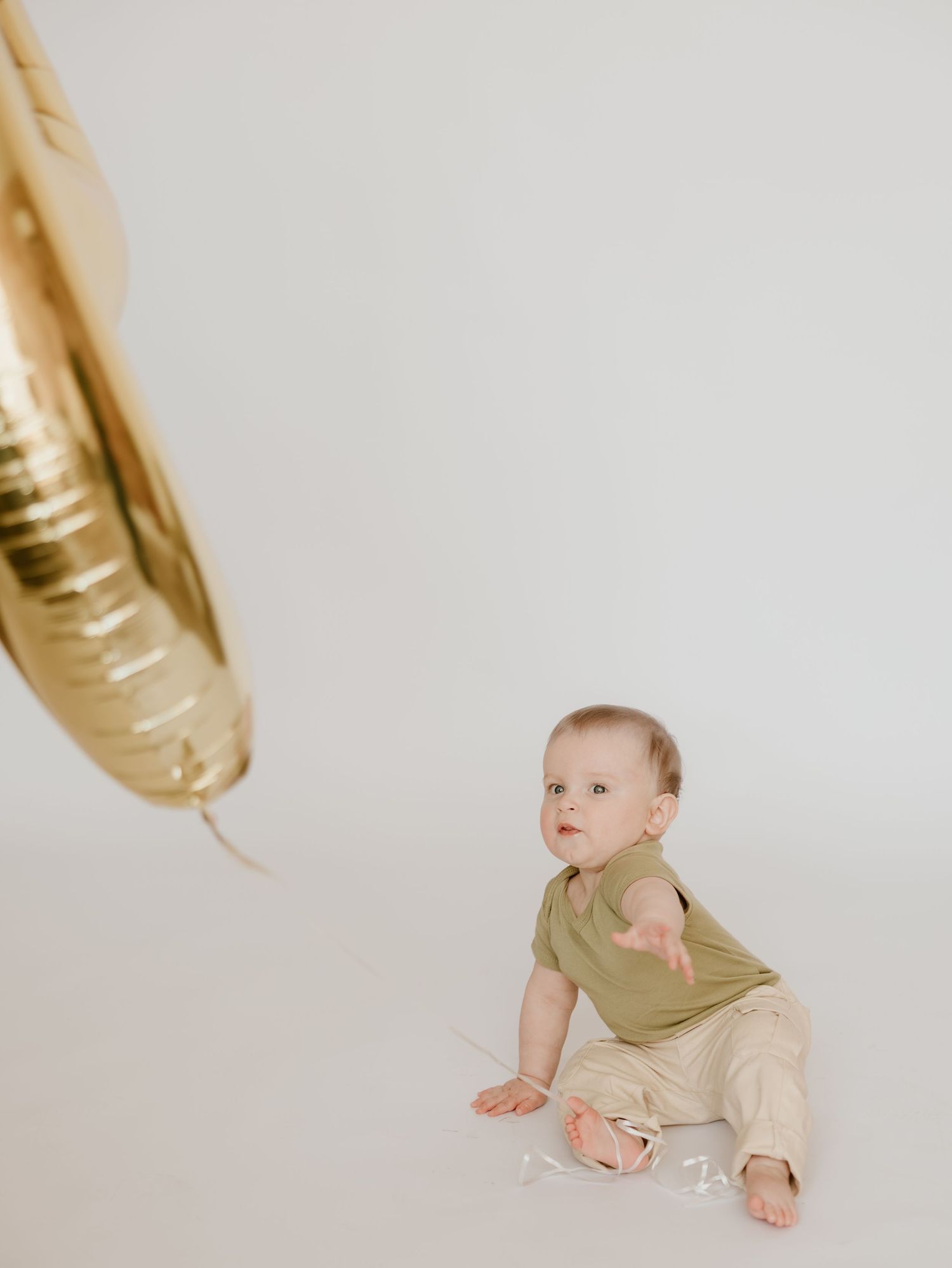 Baby in olive green shirt and beige pants sits on white floor looking up at gold balloon in minimalist setting.