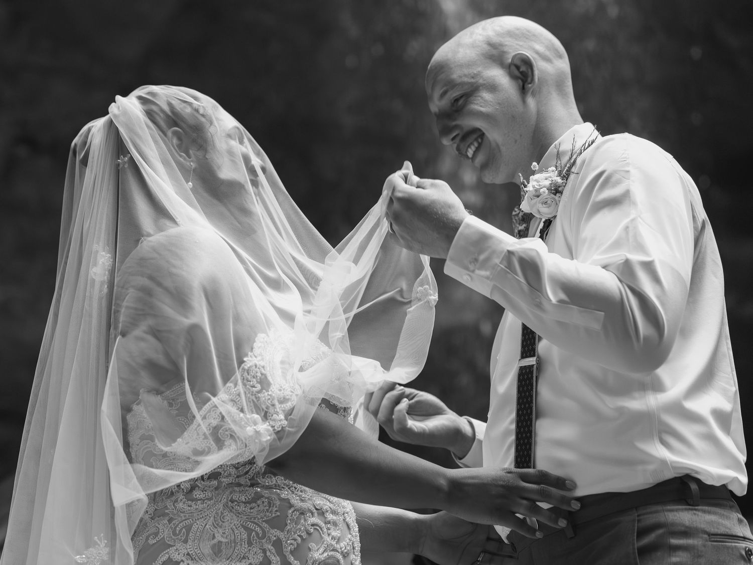A joyful black and white wedding moment showing a couple sharing laughter while the bride adjusts her veil.