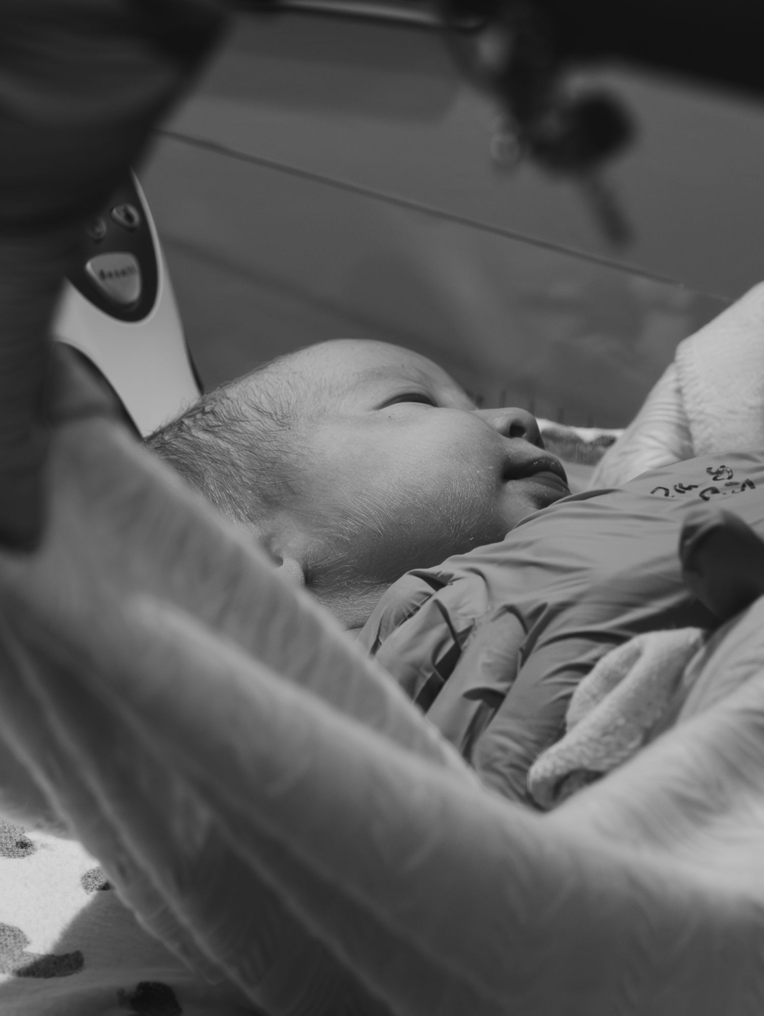 Black and white photograph shows infant laying in hospital crib while being fed from a bottle.