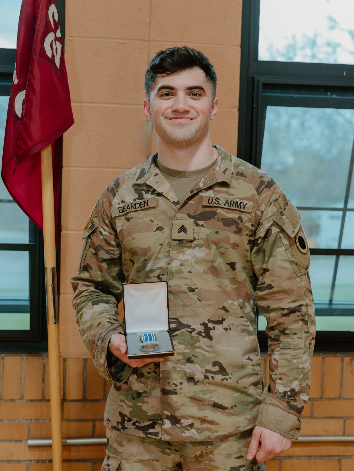 A U.S. Army service member in camouflage uniform poses with a small award or plaque against a tan wall.