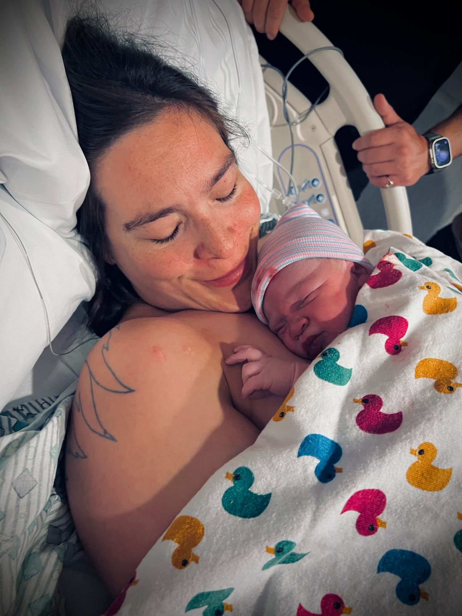 Newborn baby rests on parent's chest on hospital bed with colorful duck patterned blanket.