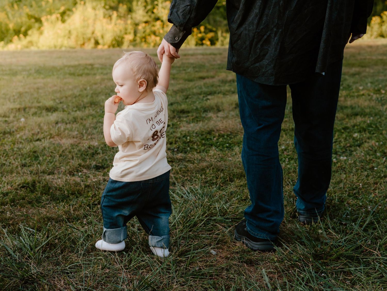 Small toddler in white outfit walking between two people's legs in grassy field.