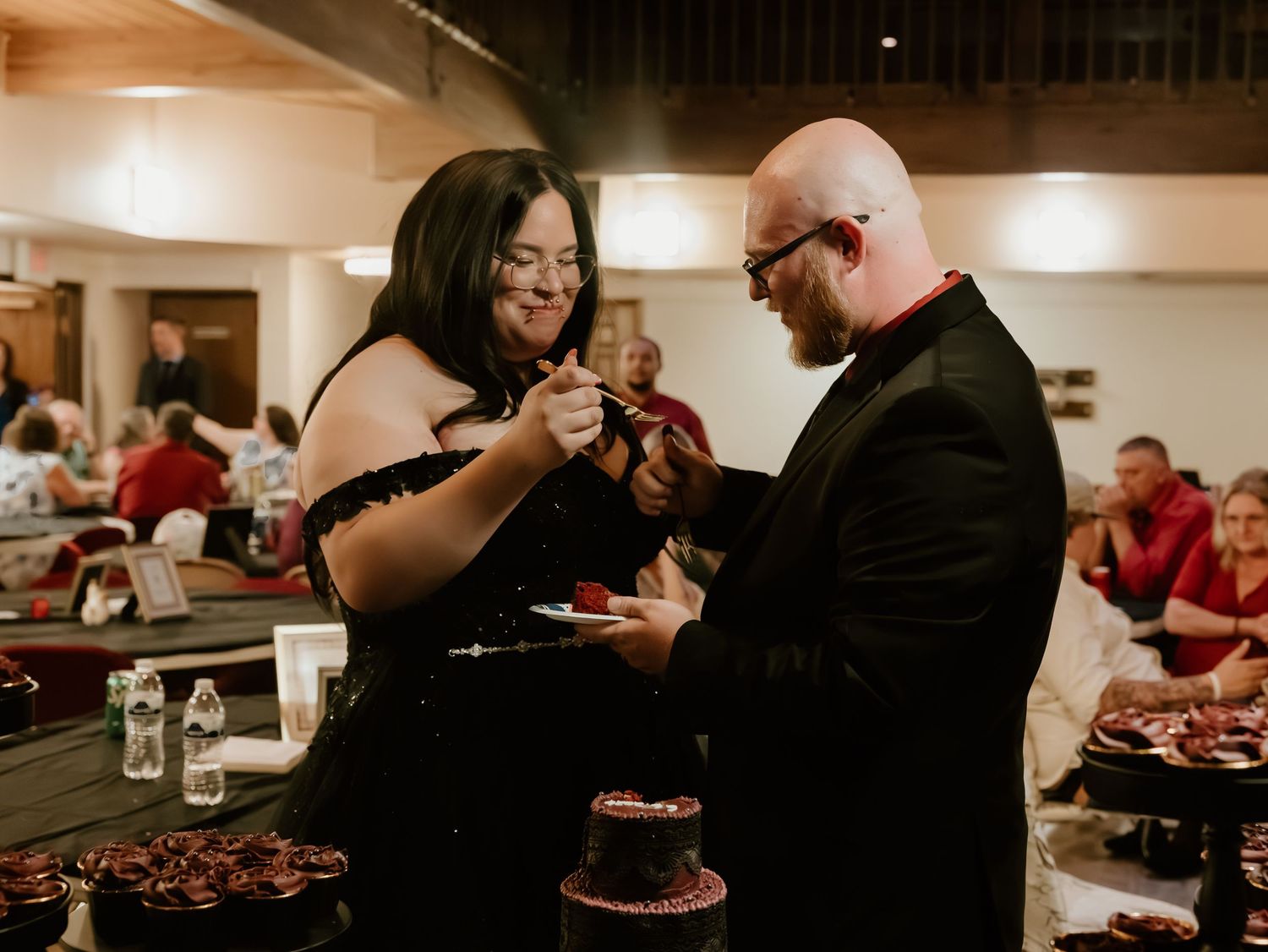 Bride and groom in black attire cutting chocolate wedding cake at elegant reception venue.