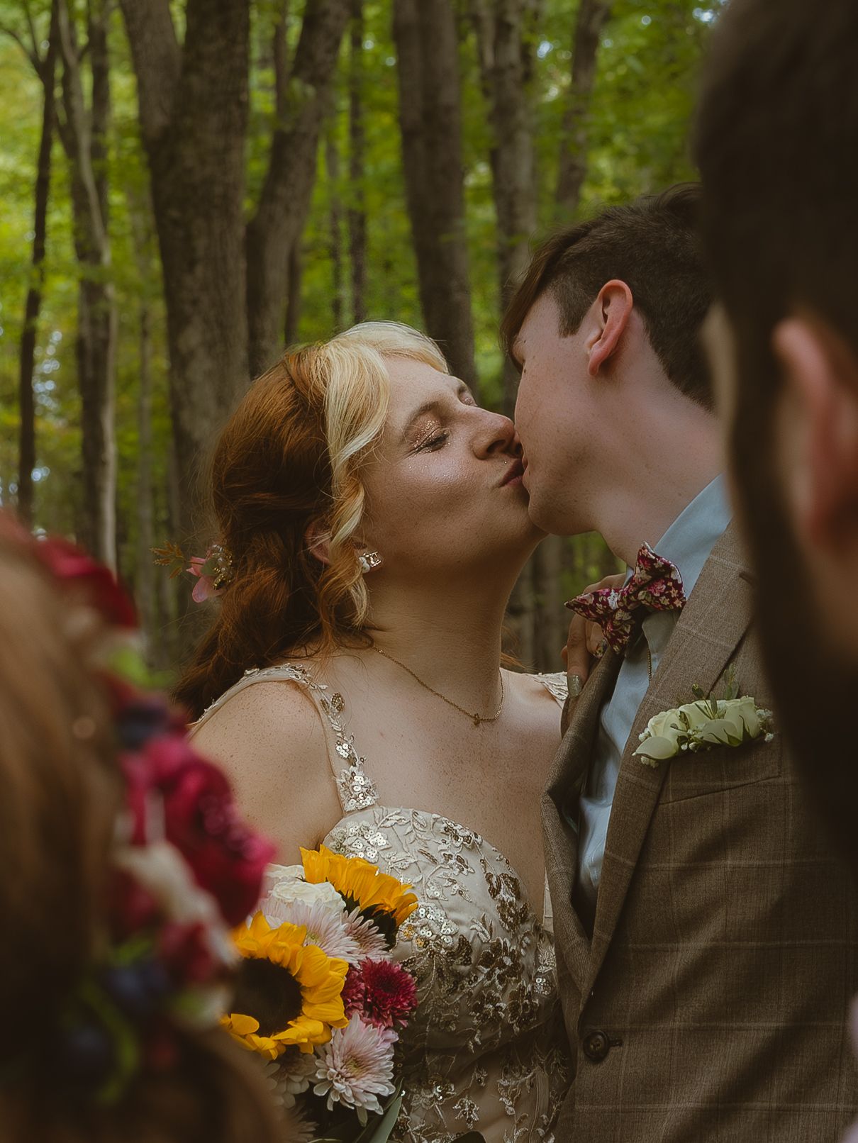 A romantic wedding couple shares a tender kiss in a sunlit forest surrounded by colorful sunflowers and burgundy florals.