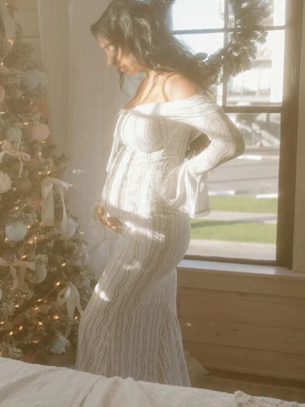 Expectant person in white lace dress poses by decorated Christmas tree near window in soft natural lighting.