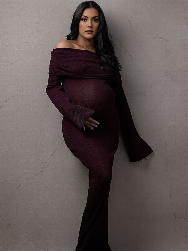 A model in a burgundy off-shoulder knit dress poses against a textured gray wall in a maternity-style photoshoot.