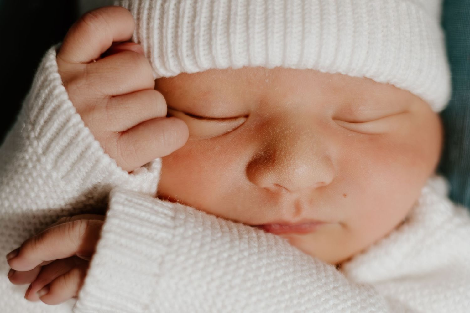 Sleeping newborn baby wearing white knit hat peacefully rests with tiny hand near face.