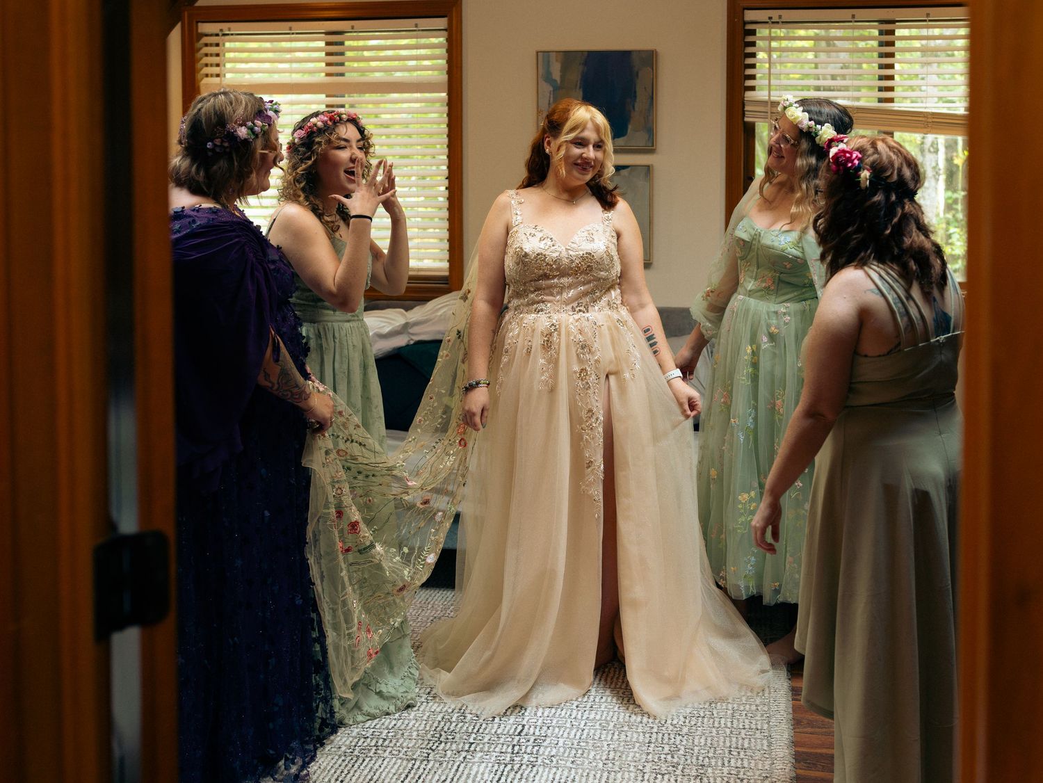 A group of bridesmaids in pastel sage green dresses help the bride get ready in a sunlit room.