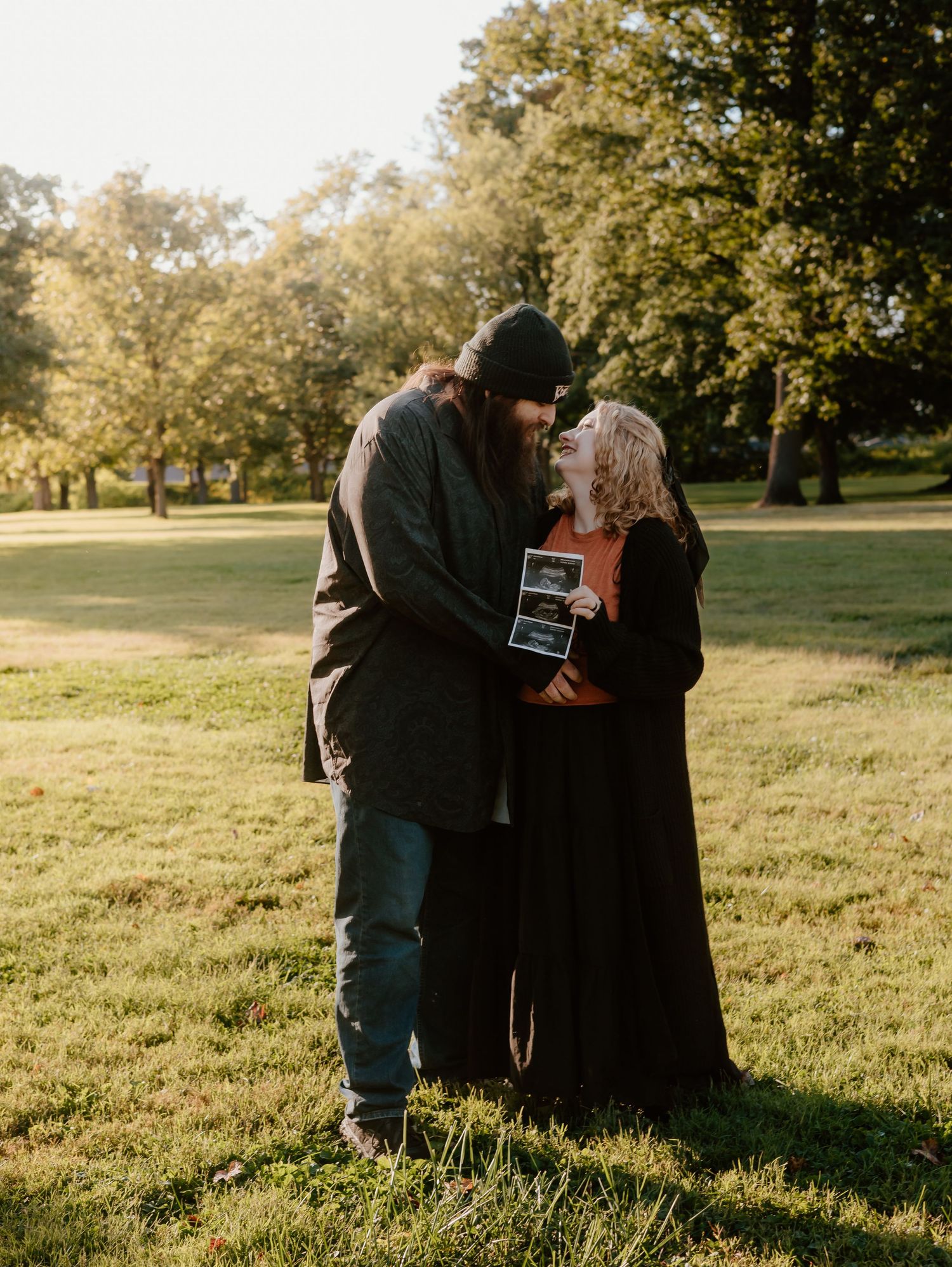 Couple holding ultrasound photos while embracing in a sunlit park during autumn.