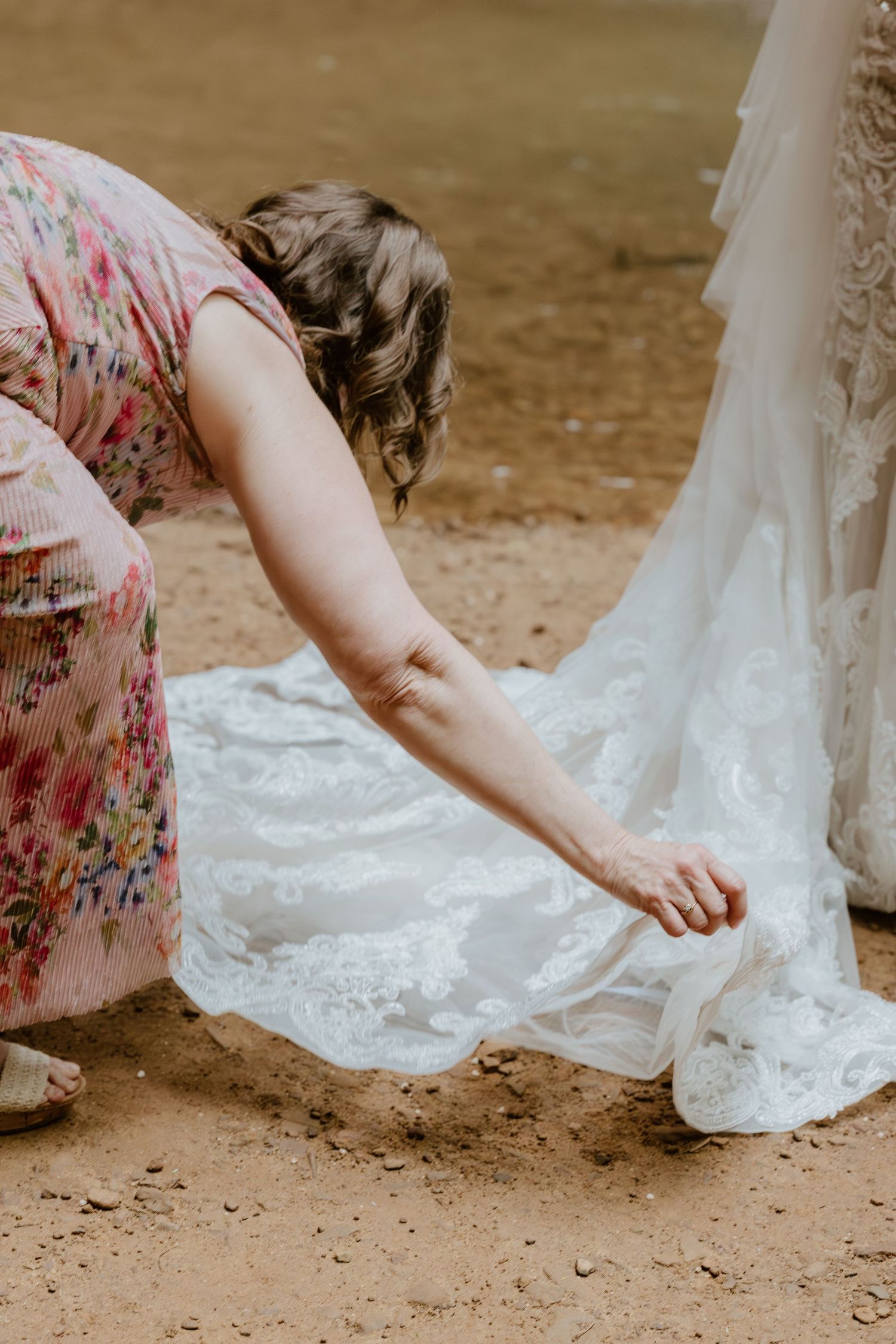 Close-up view of hands holding the hem of a white wedding dress on dirt ground.