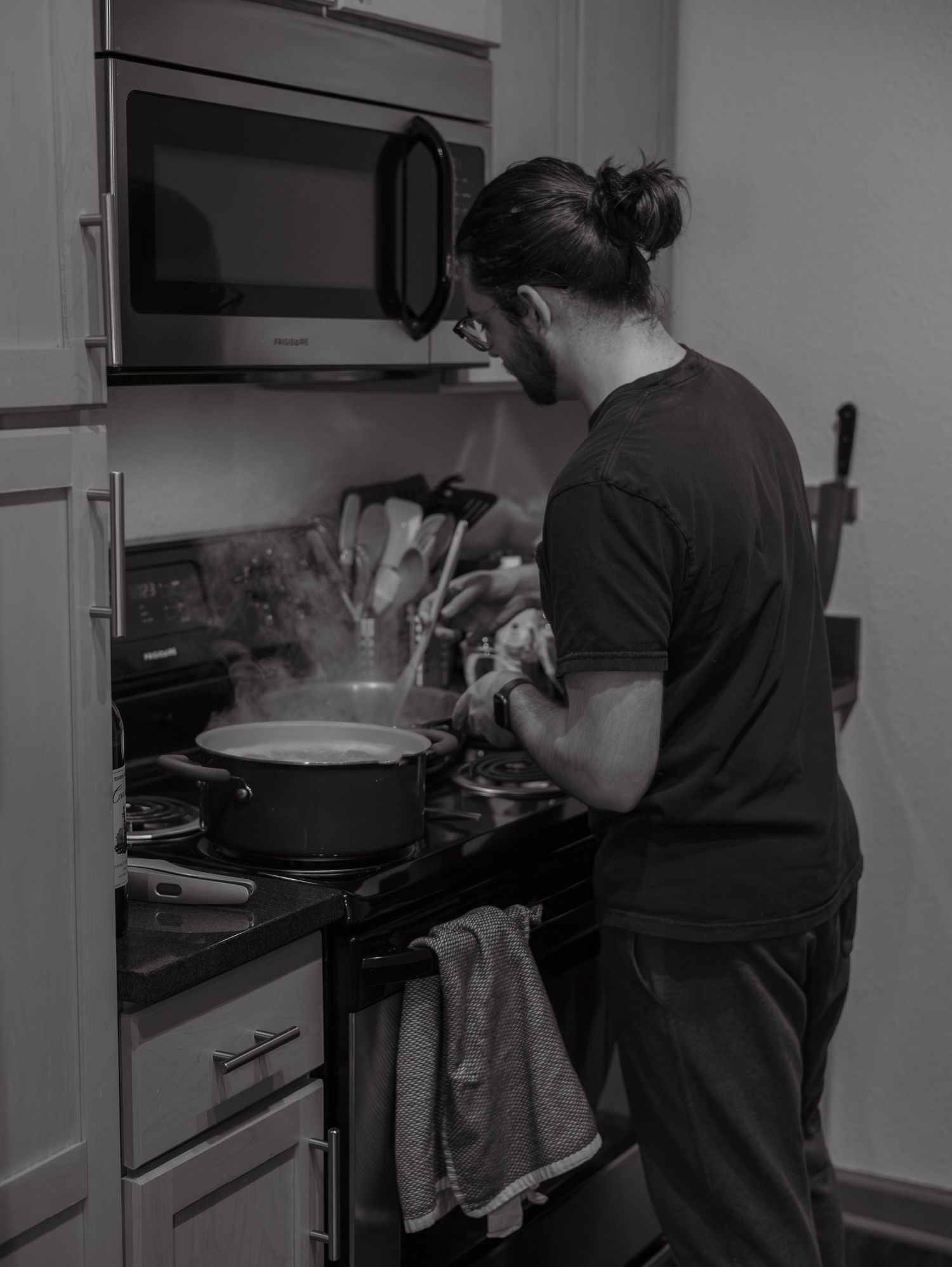 A person in dark clothing stands at a stovetop cooking in a kitchen with a microwave mounted above.