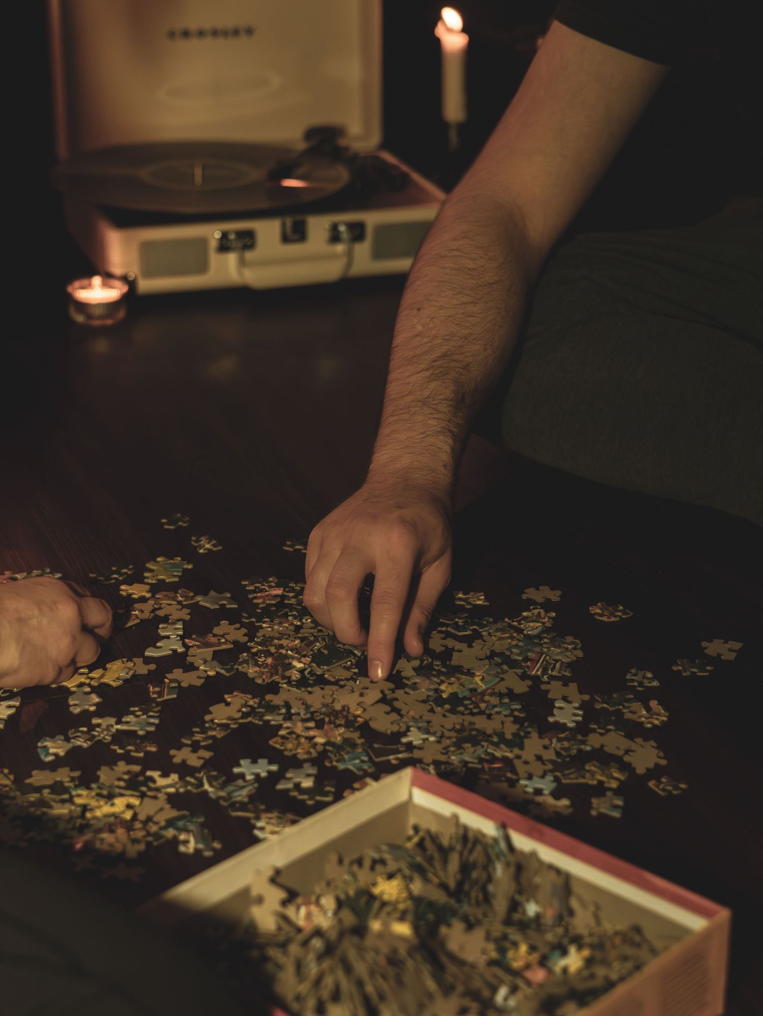 Hands work on assembling a jigsaw puzzle by candlelight on a dark wooden table in a dimly lit room.