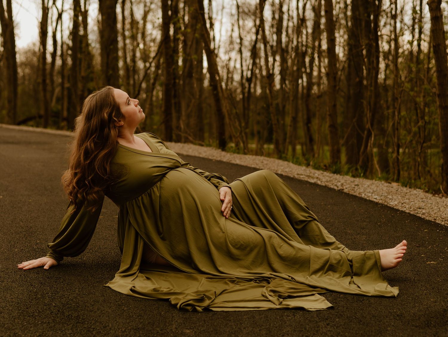 Pregnant woman in olive green gown reclines on forest road, long red hair flowing, barefoot among autumn trees.