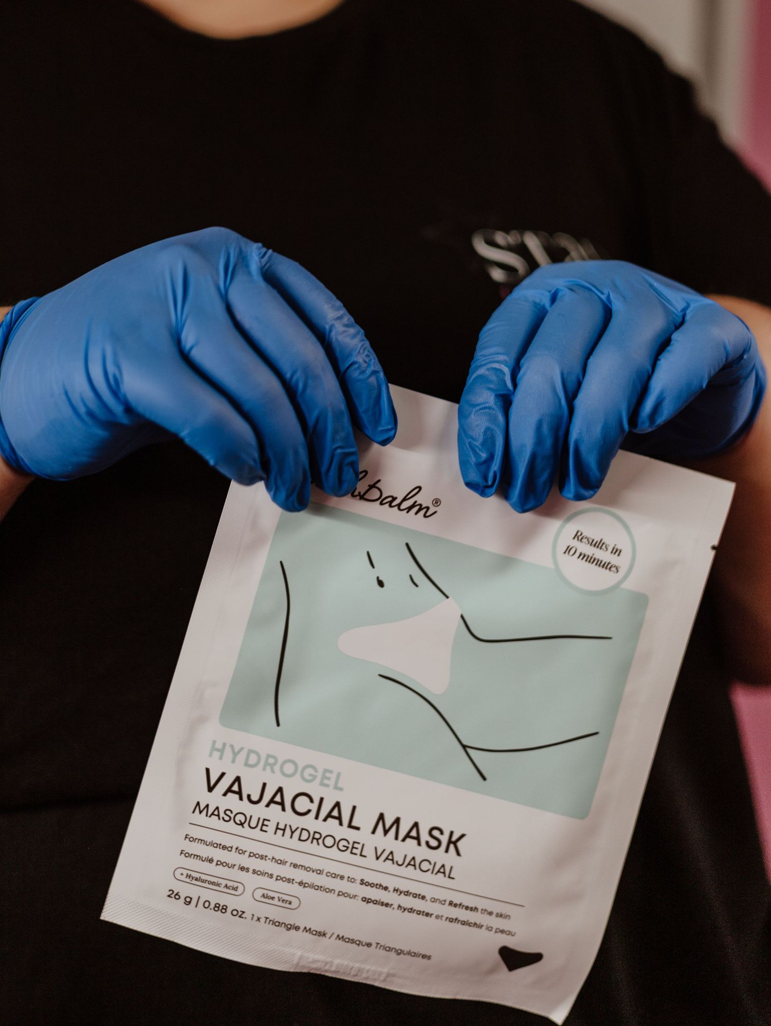 Hands wearing blue medical gloves hold up a white facial mask packet with minimalist design against dark background.