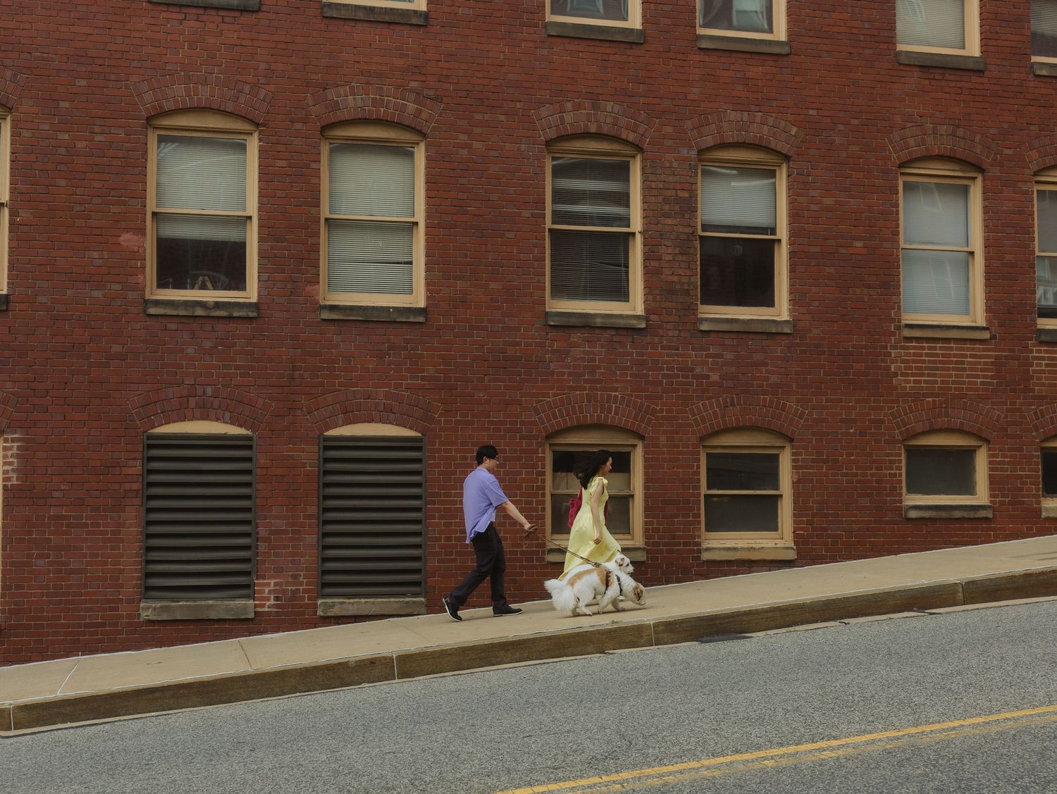 Couple in formal attire walking past a large red brick building on a city sidewalk.